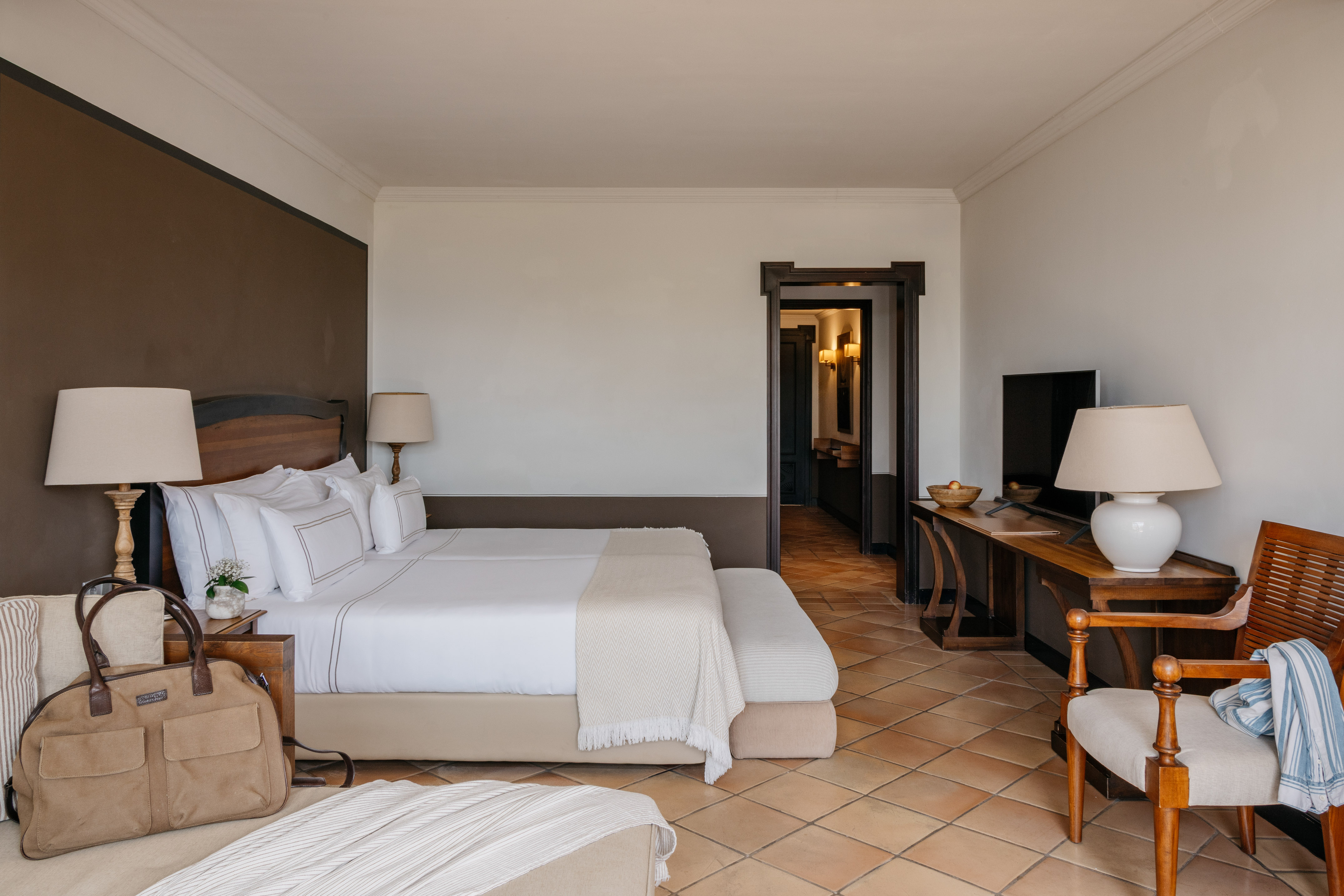 Luxury hotel bedroom with king bed, white linens, wood furniture, terracotta floor.