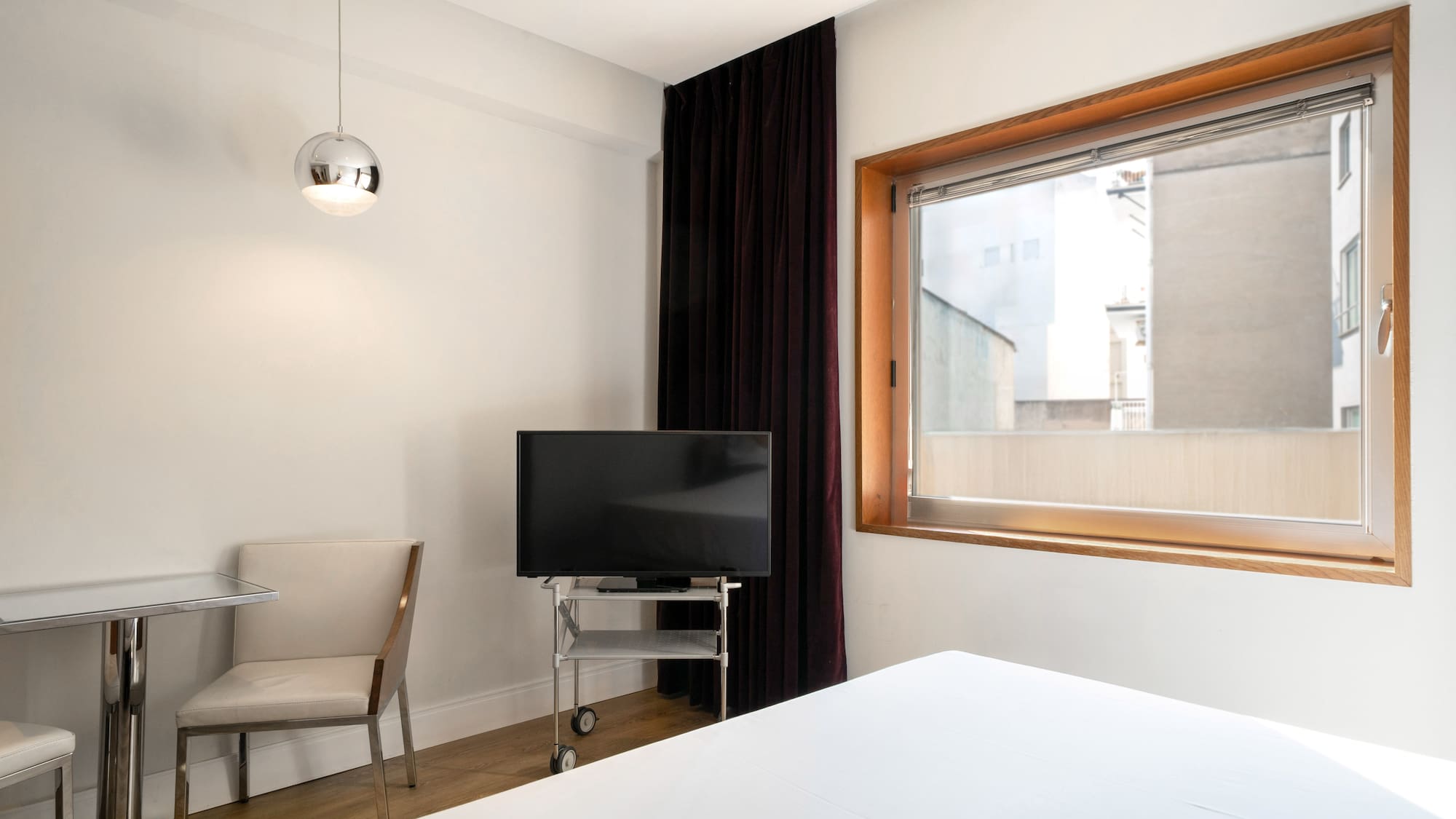 Modern hotel room with white bed, TV on wheels, wood window, small table.