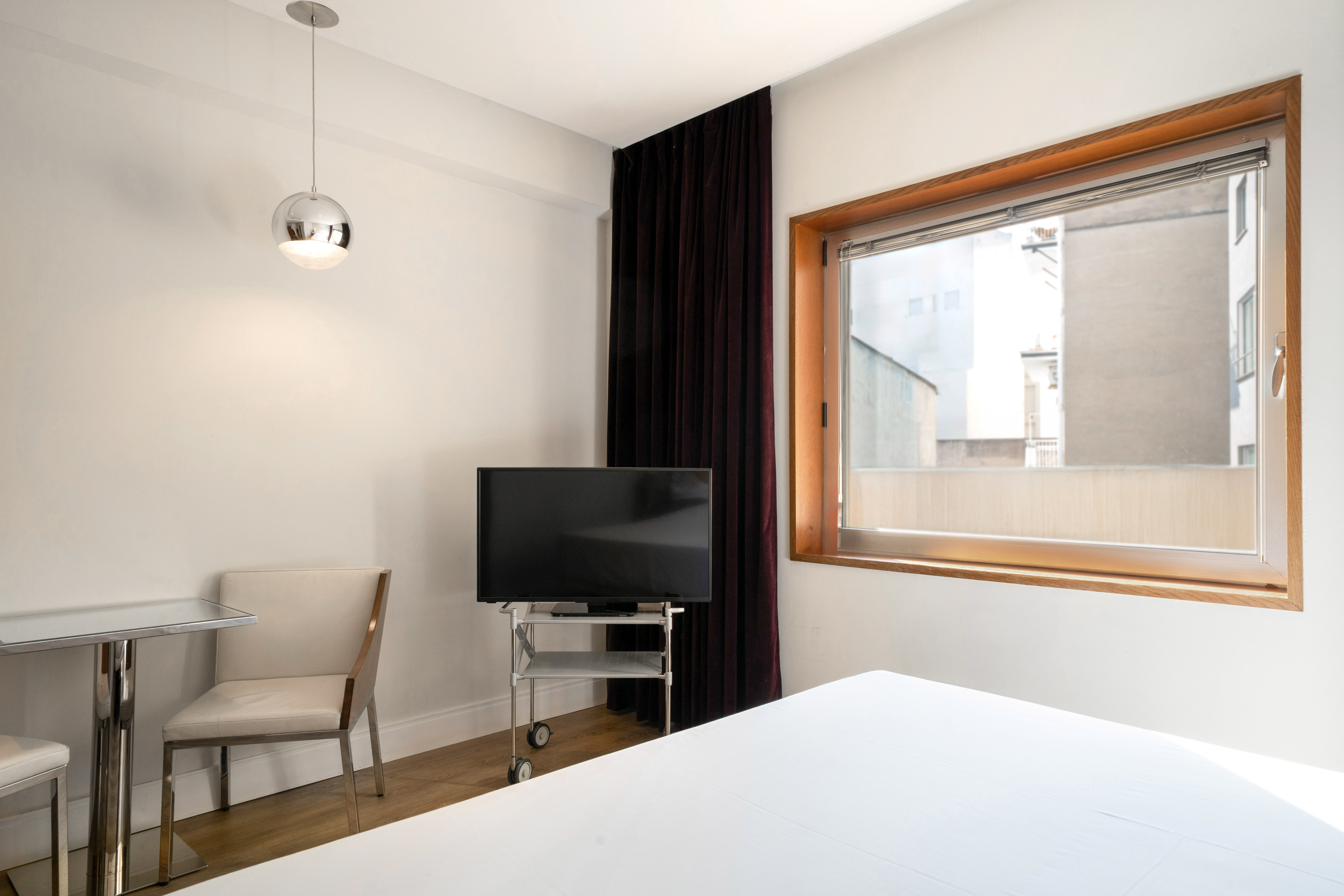 Modern hotel room with white bed, TV on wheels, wood window, small table.