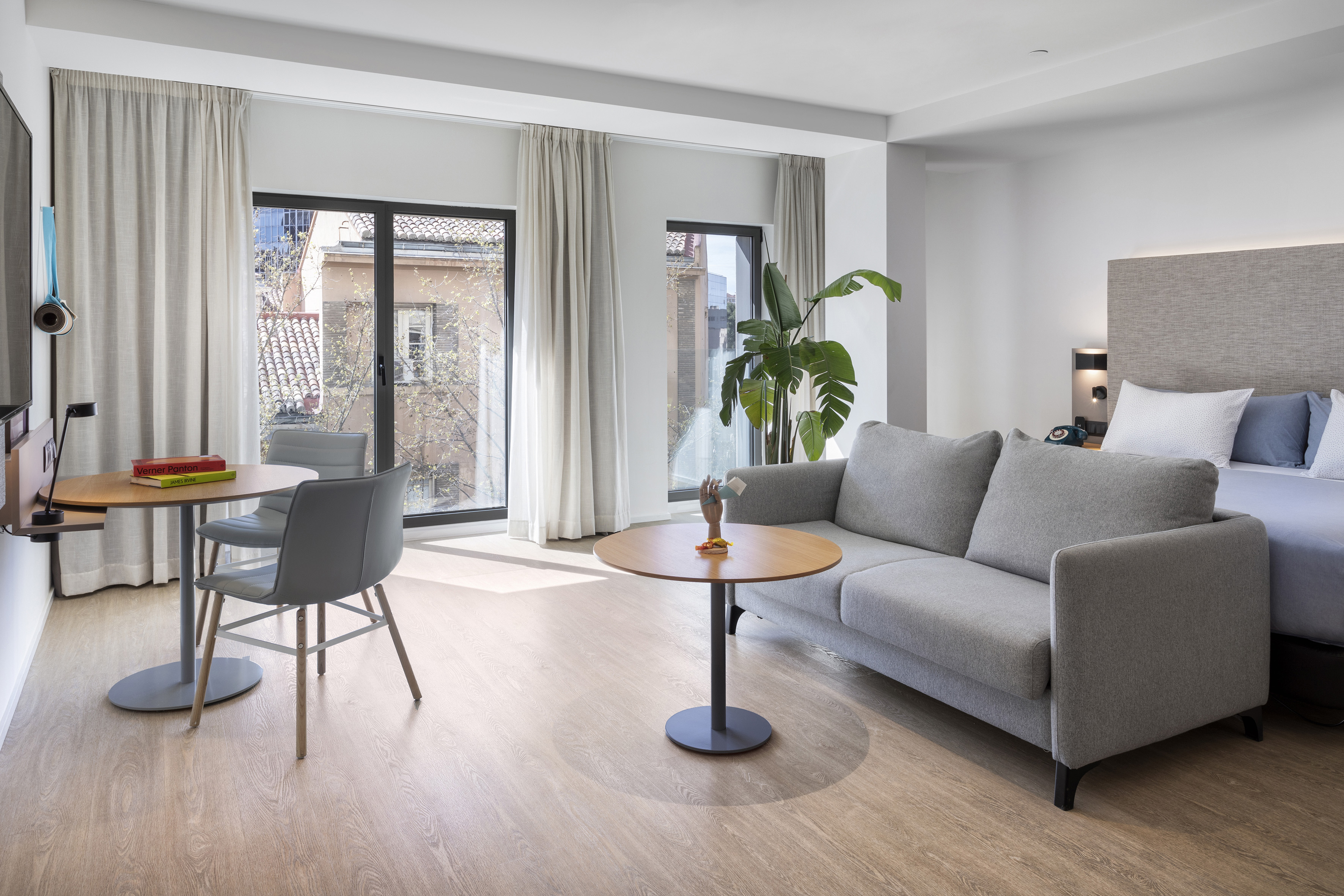 Modern hotel studio suite, grey sofa, bed, work table, bright windows.