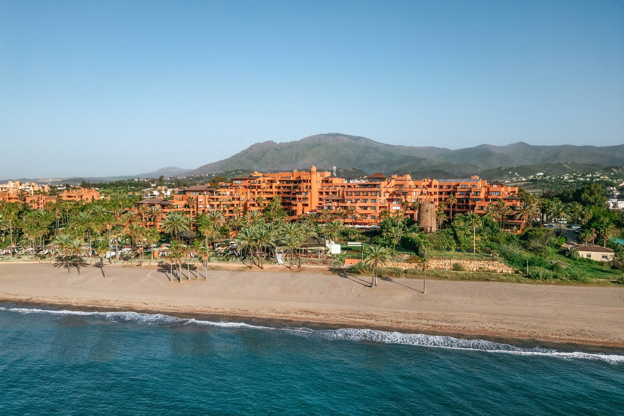 Beachfront hotel with orange buildings, palm trees, sandy beach, and blue sea.