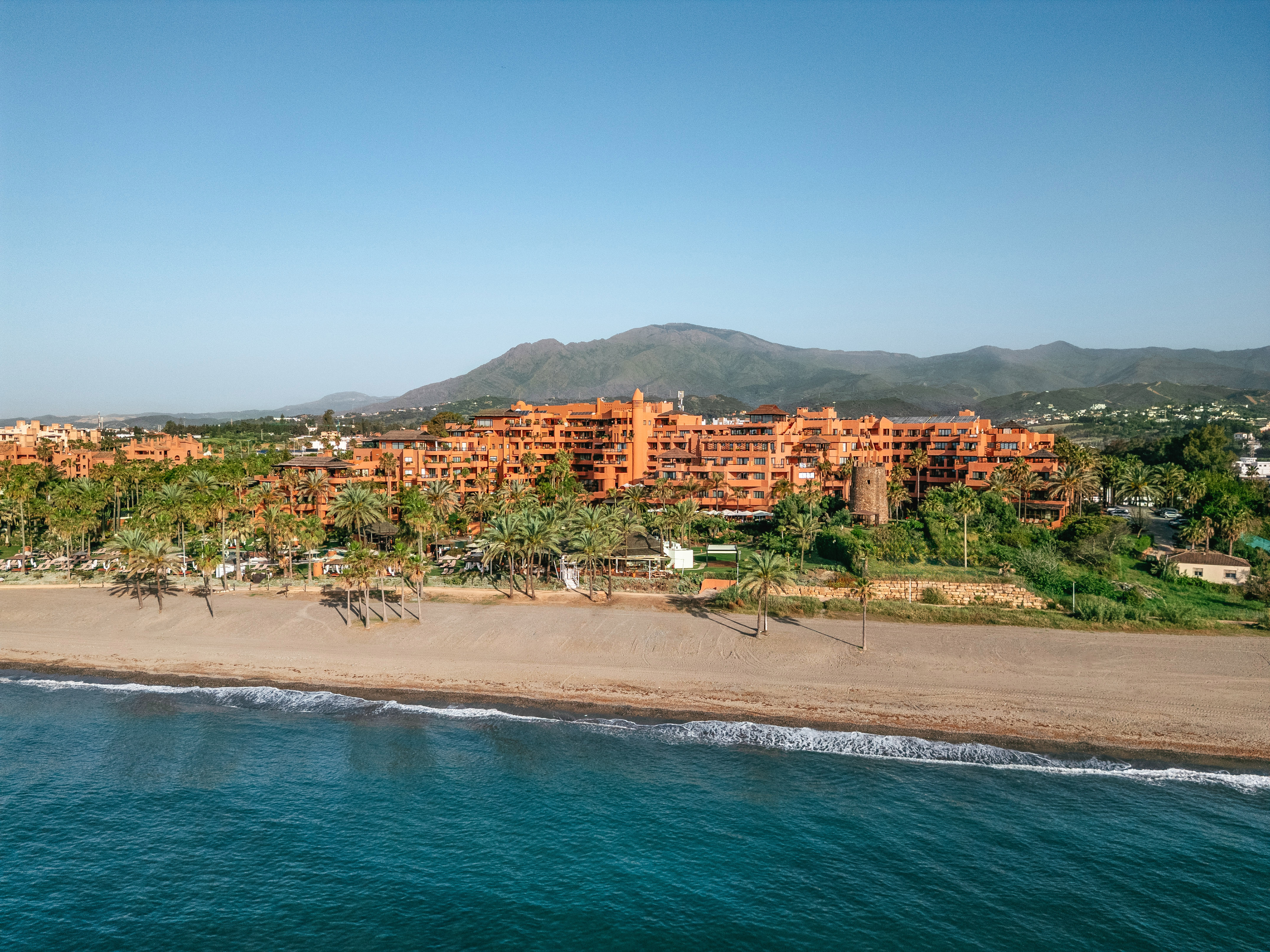Beachfront hotel with orange buildings, palm trees, sandy beach, and blue sea.