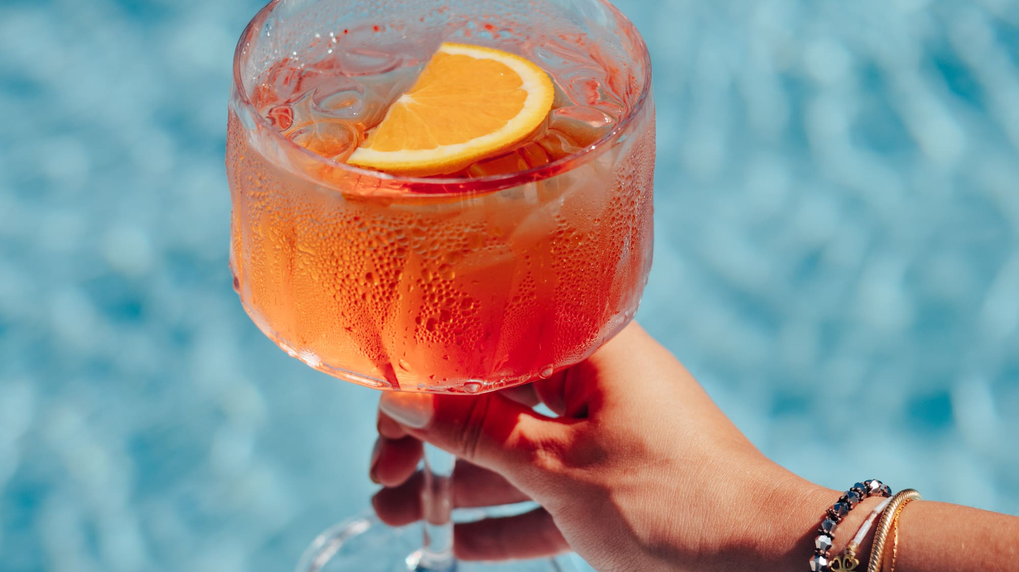 Hand holding refreshing orange spritz cocktail with orange slice over blue pool.
