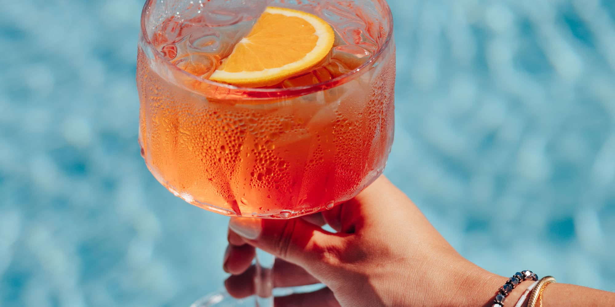 Hand holding refreshing orange spritz cocktail with orange slice over blue pool.
