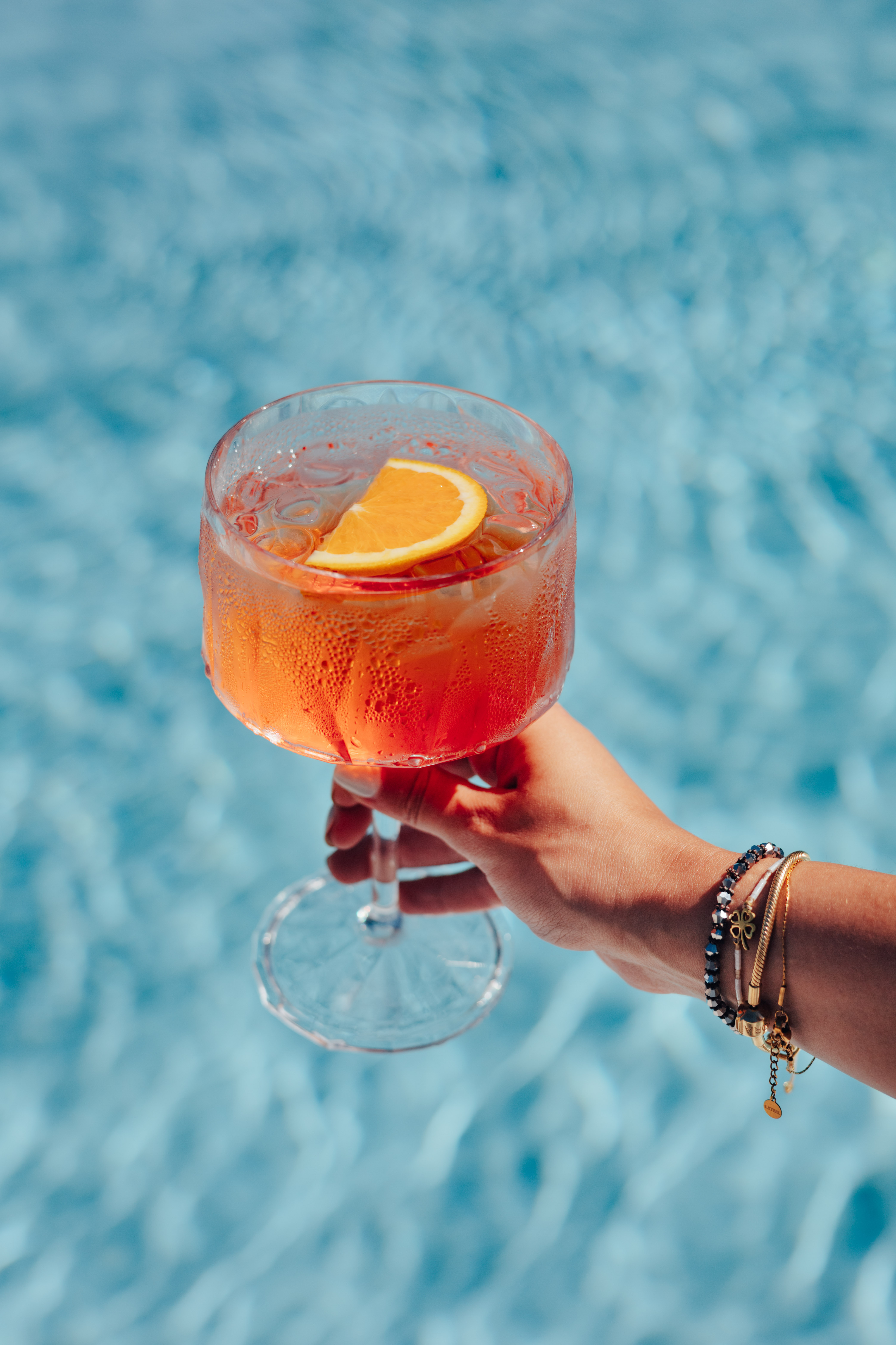 Hand holding refreshing orange spritz cocktail with orange slice over blue pool.
