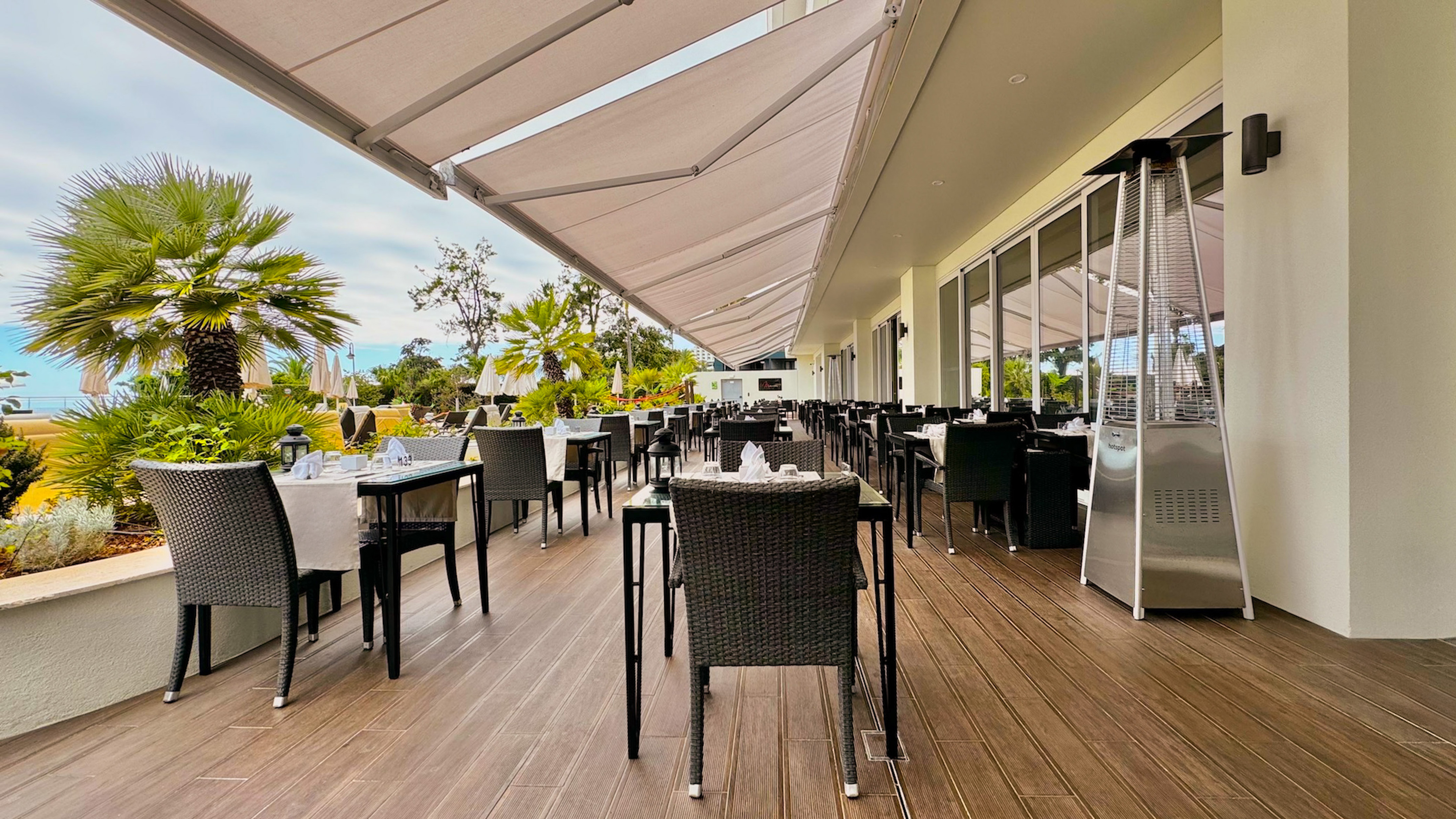 Resort outdoor dining terrace with tables, retractable awnings, and tropical palm trees.
