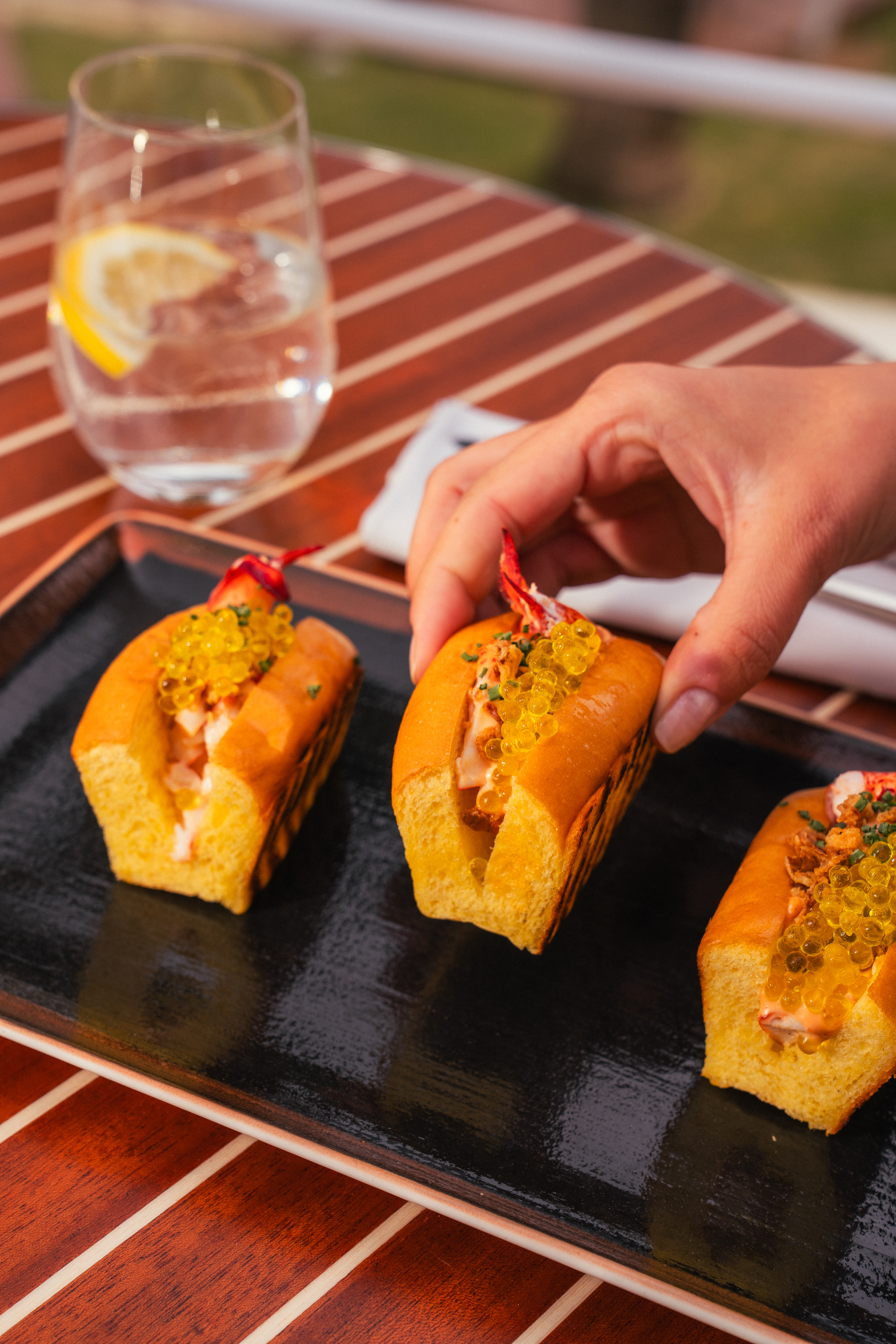 Hand picking a brioche lobster roll with golden caviar from a black tray.