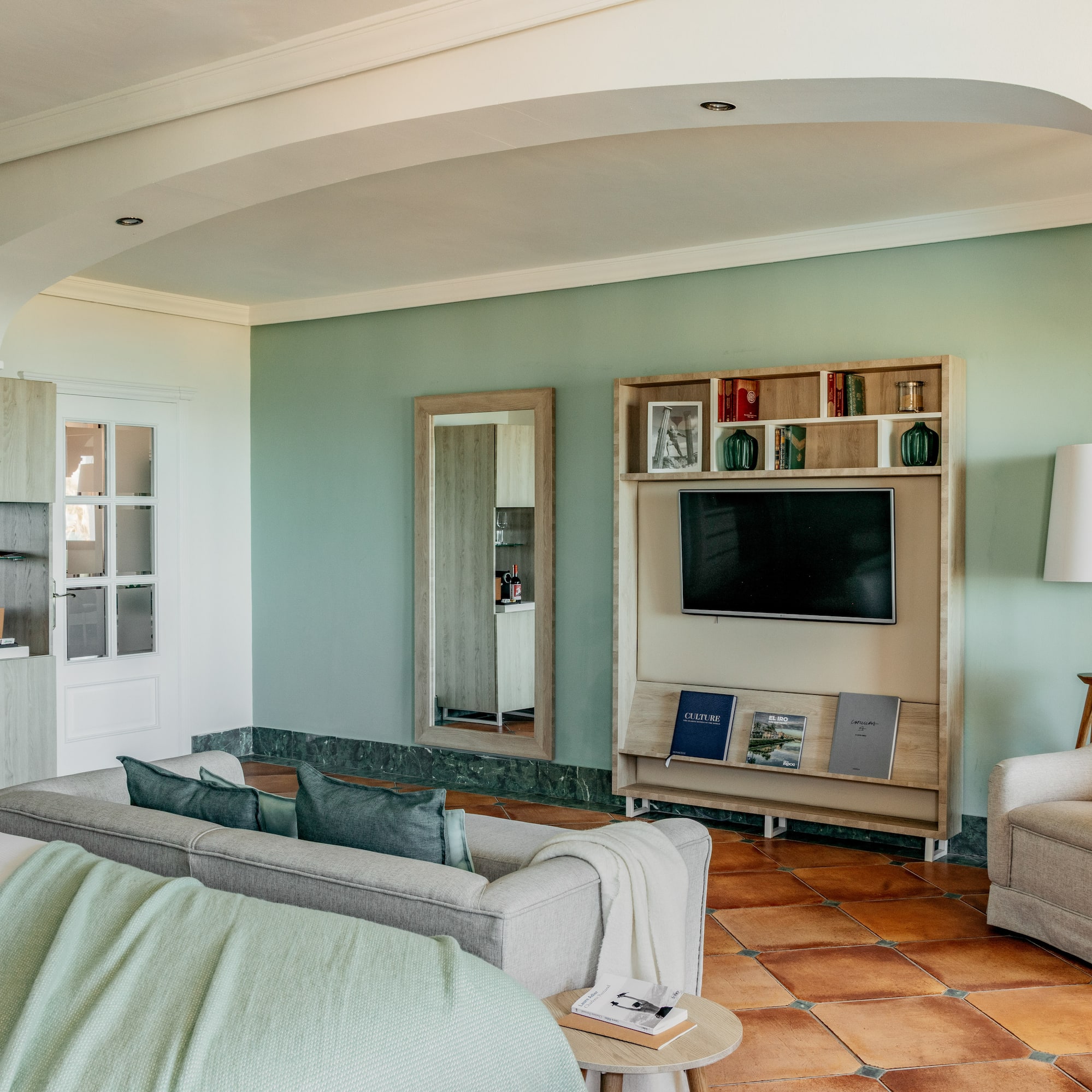 Hotel living room with green walls, gray sofa, TV, and terracotta floor.