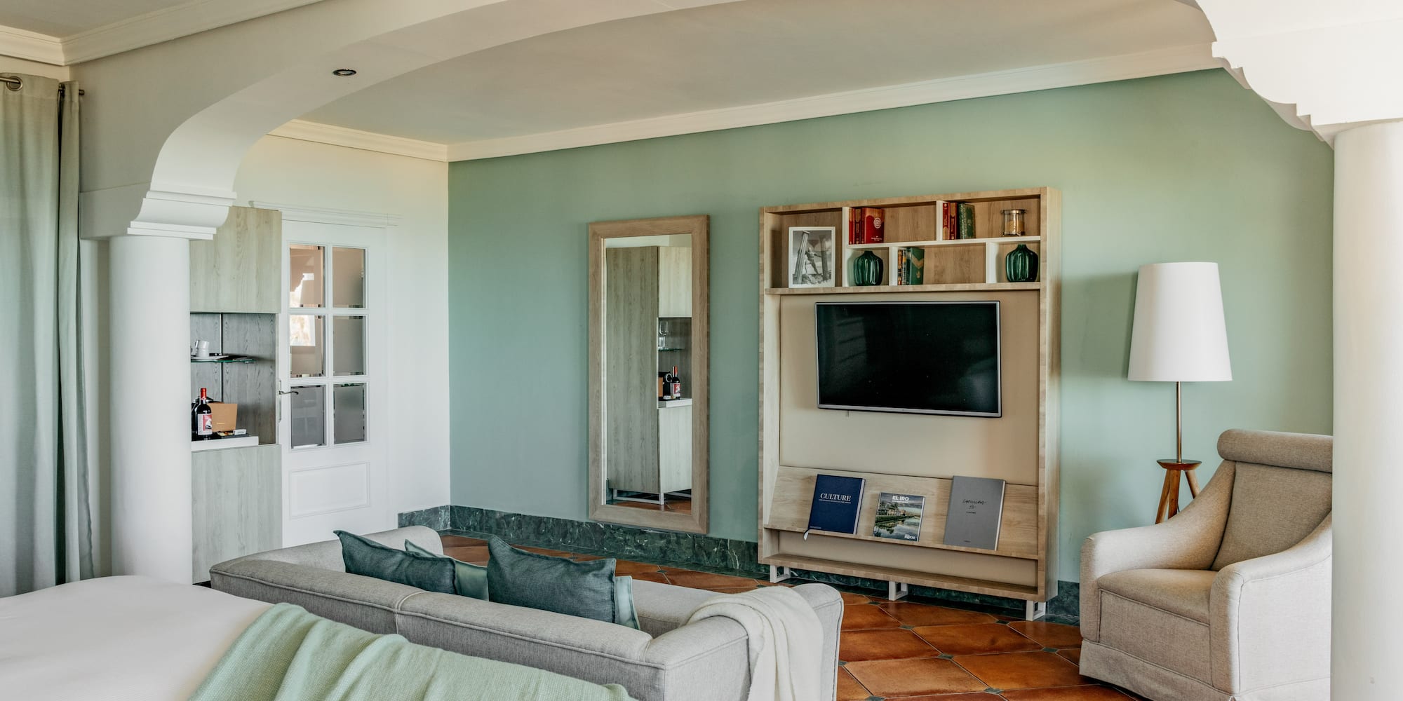 Hotel living room with green walls, gray sofa, TV, and terracotta floor.