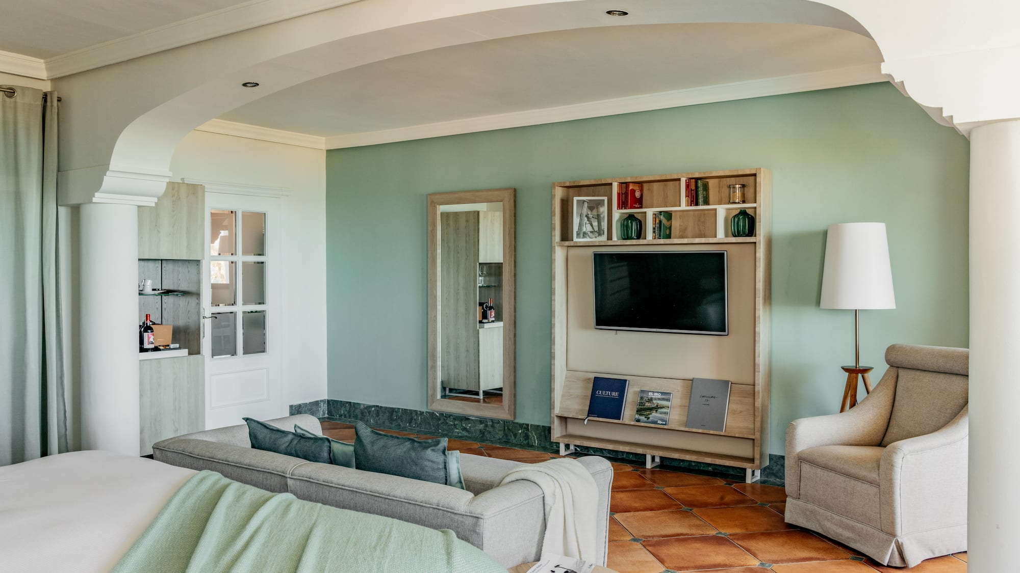 Hotel living room with green walls, gray sofa, TV, and terracotta floor.
