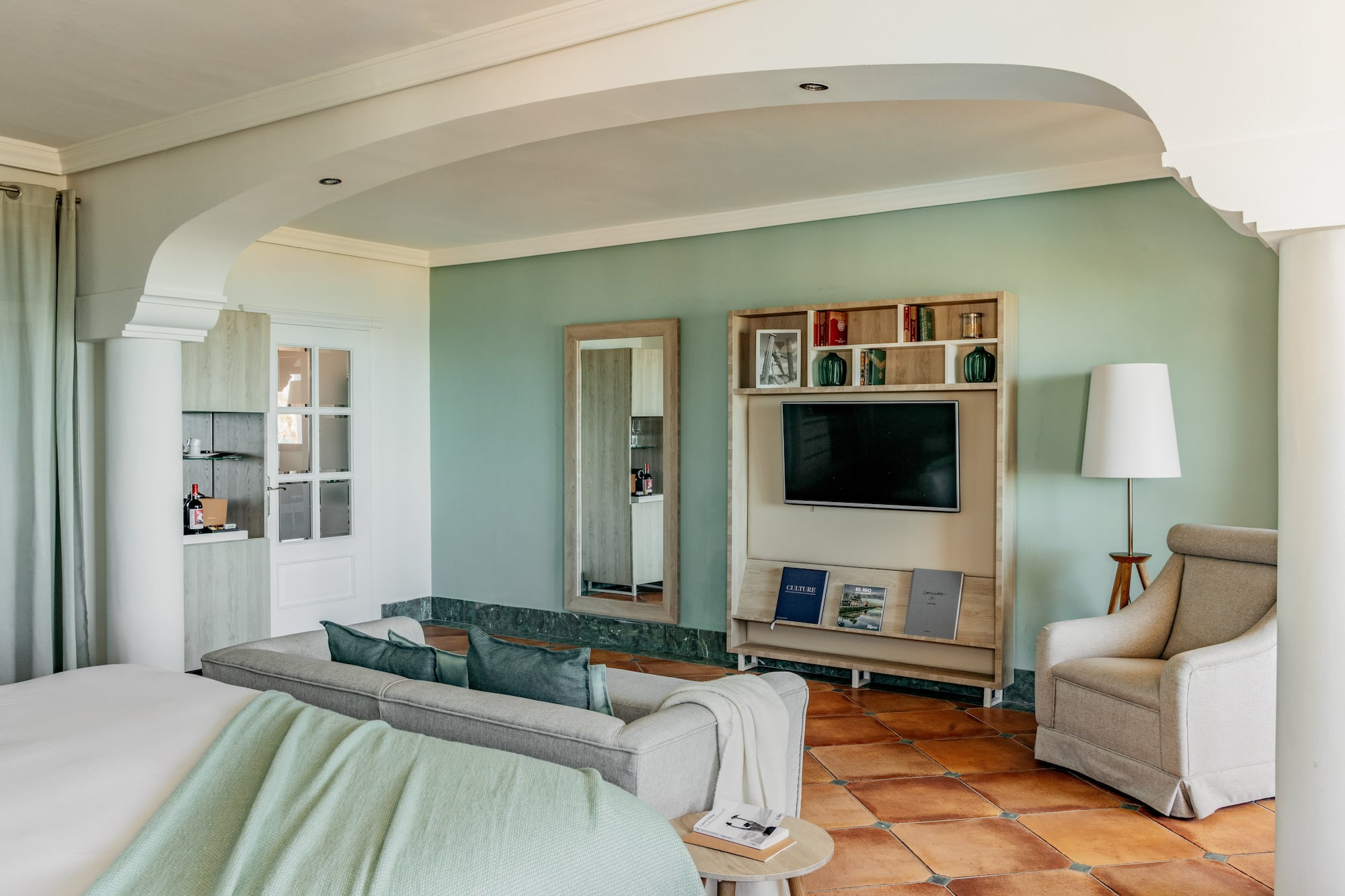 Hotel living room with green walls, gray sofa, TV, and terracotta floor.