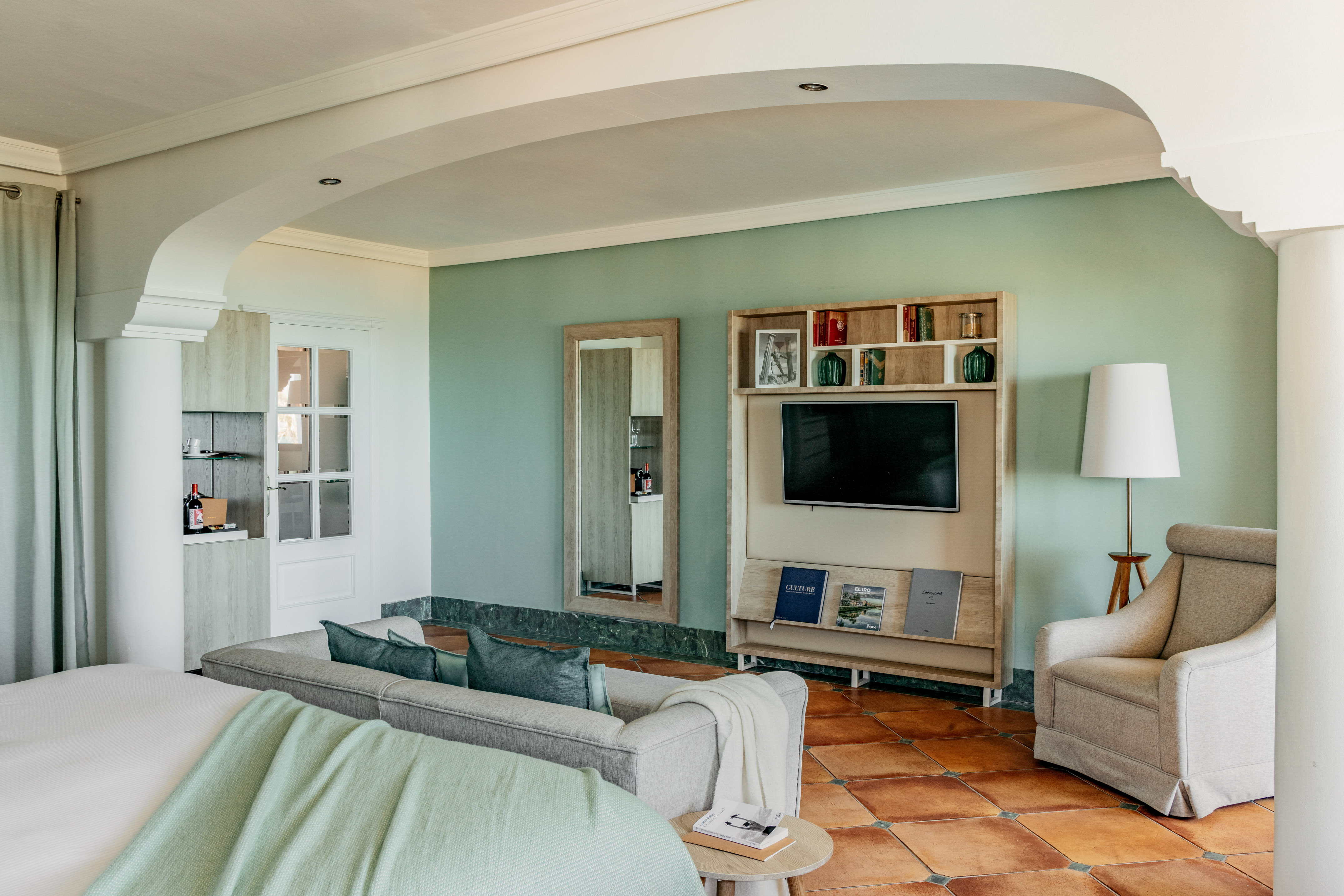 Hotel living room with green walls, gray sofa, TV, and terracotta floor.
