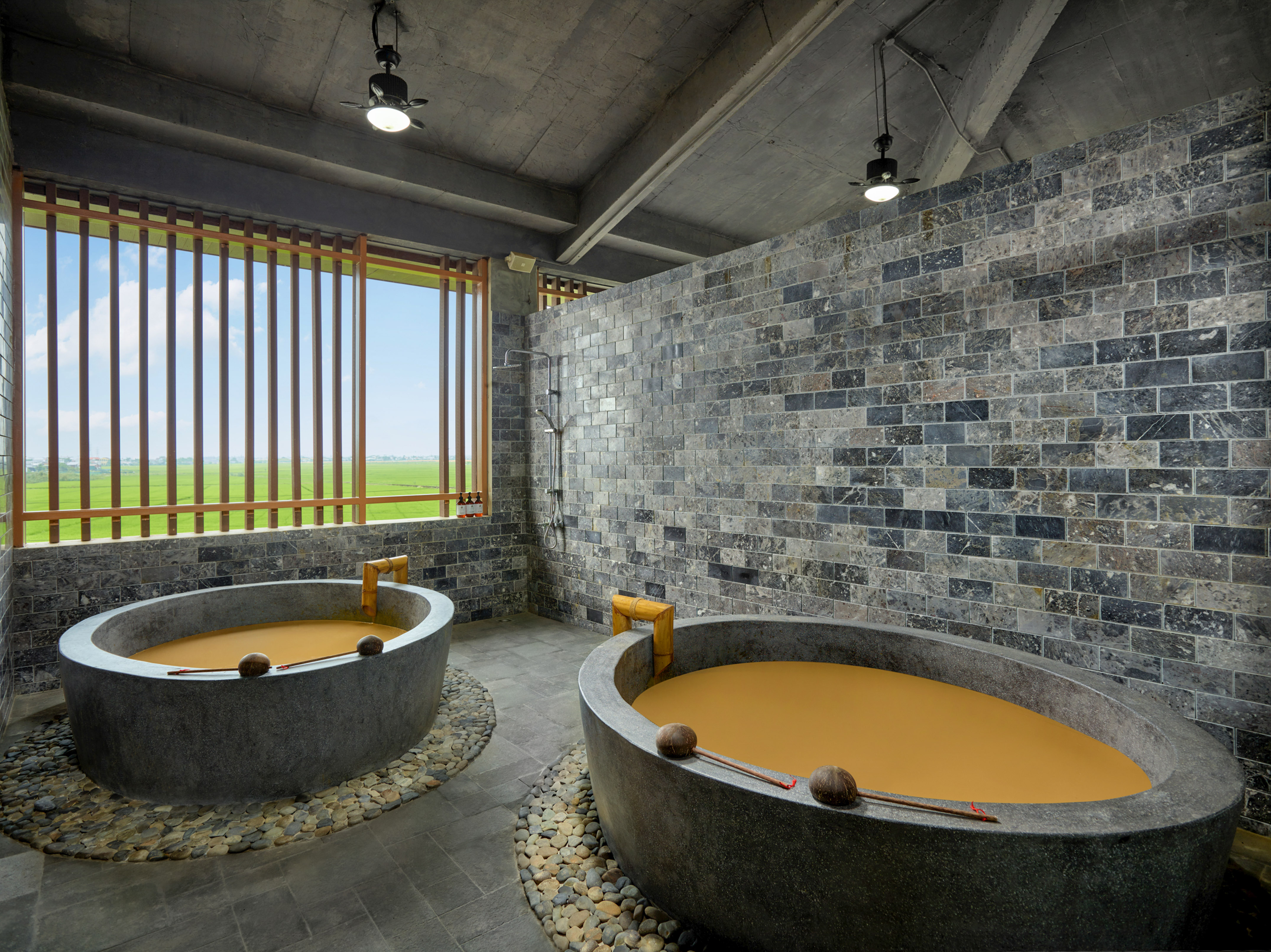 Rustic spa with two stone mud bathtubs, tile walls, and rice field view.