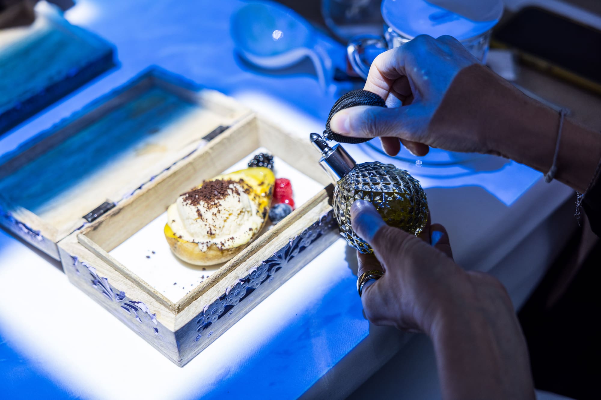 Hands spraying perfume on gourmet ice cream dessert with berries in a box.