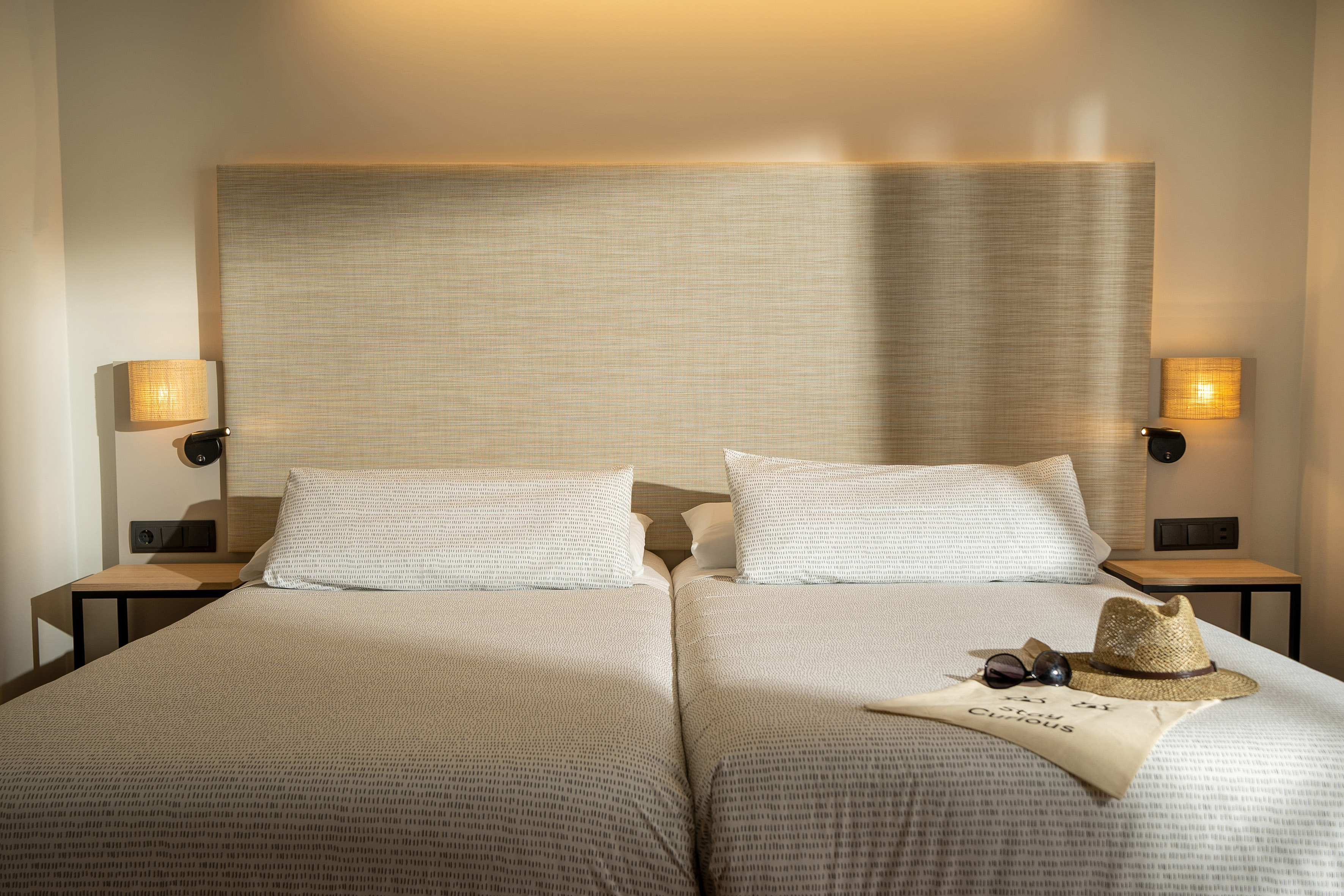 Hotel room with twin beds, woven headboard, bedside lamps, straw hat, sunglasses.