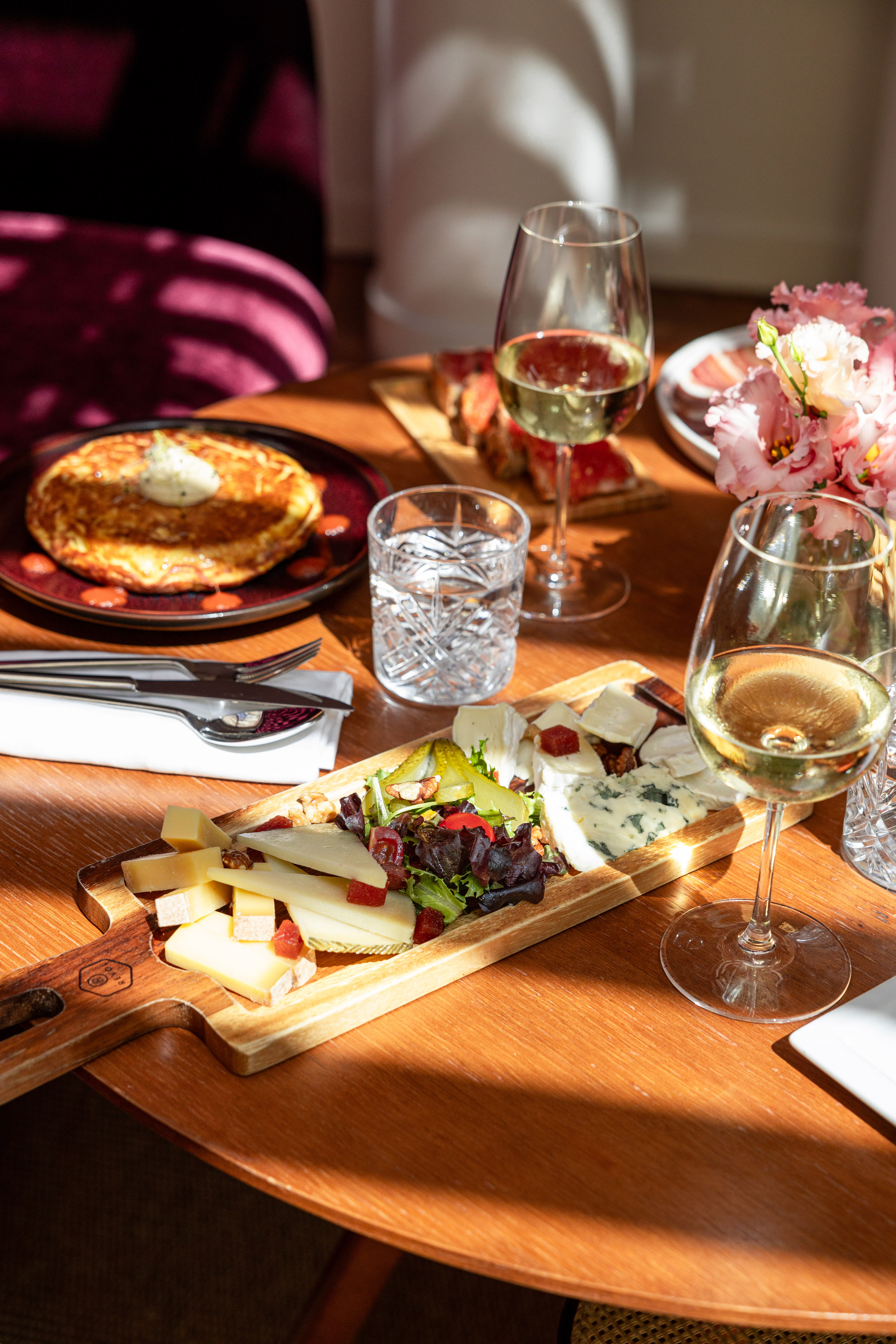 Elegant cheese board, white wine, and omelet on a sunlit wooden table.