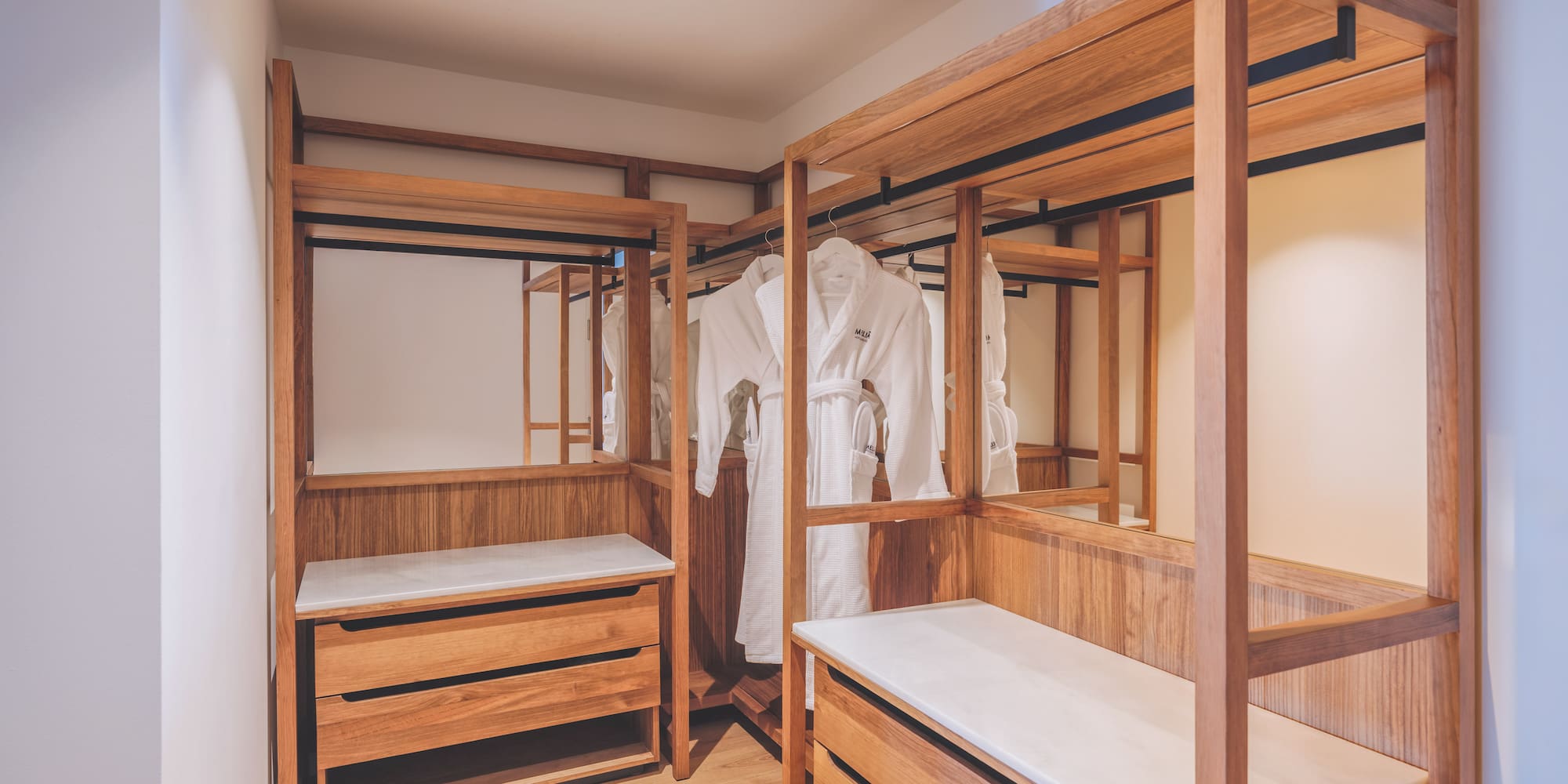 Wood walk-in closet with white bathrobes, storage drawers, and mirrors.