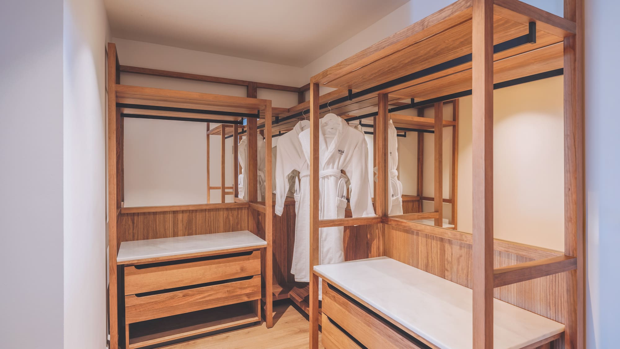 Wood walk-in closet with white bathrobes, storage drawers, and mirrors.