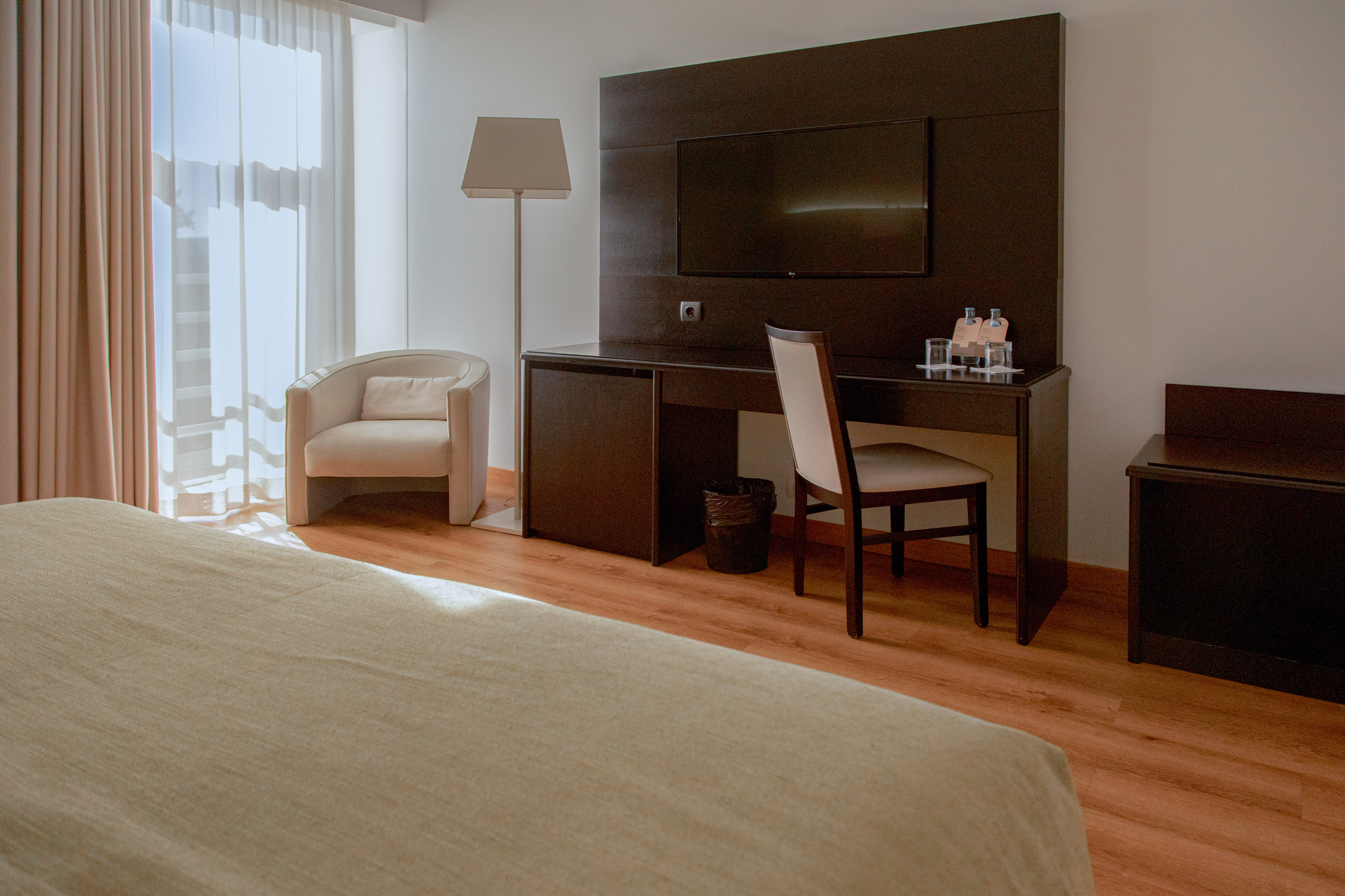 Modern hotel room with desk, TV, armchair, and sheer window curtains.