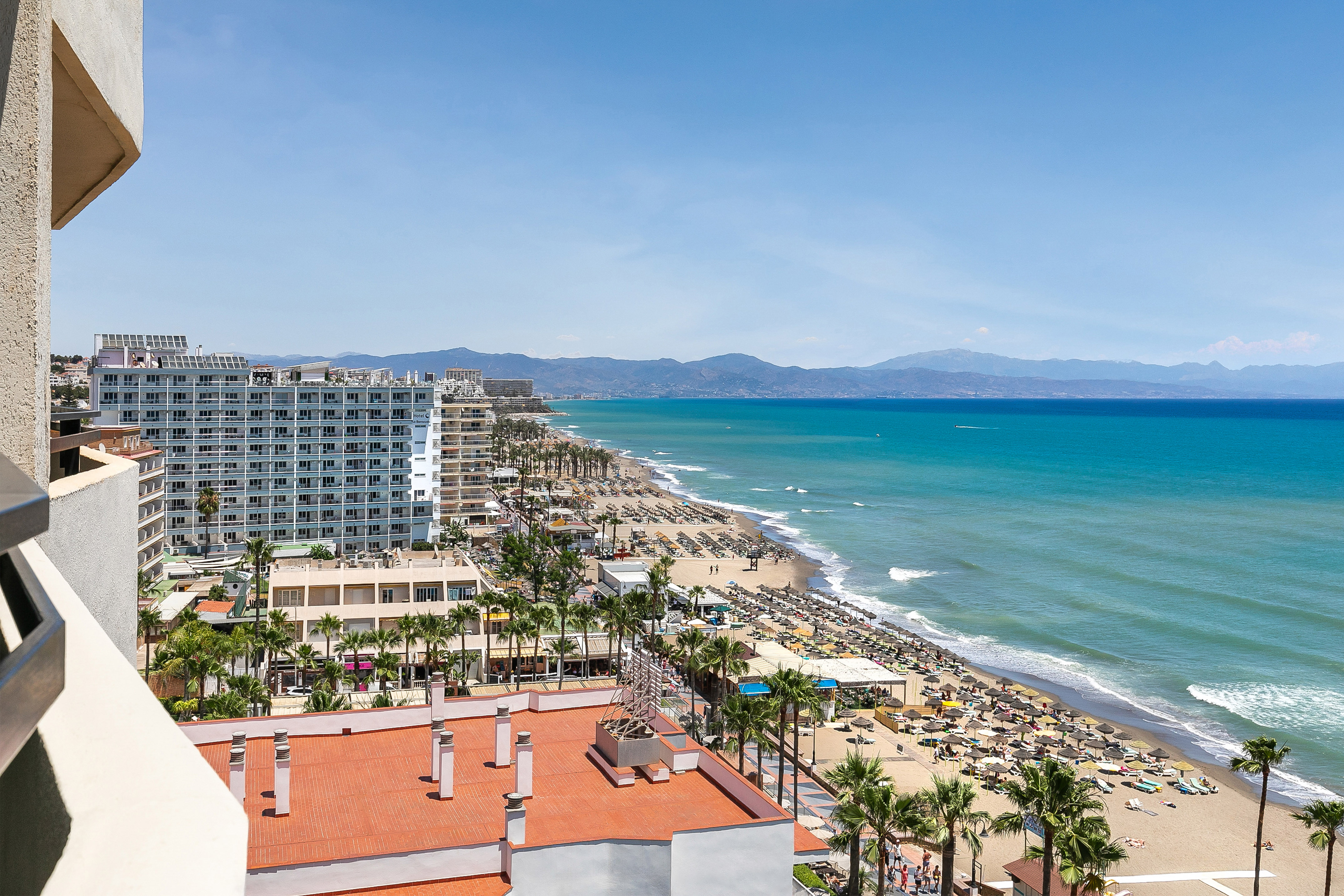 Sunny beach resort with hotels, palm trees, turquoise sea, and mountains.
