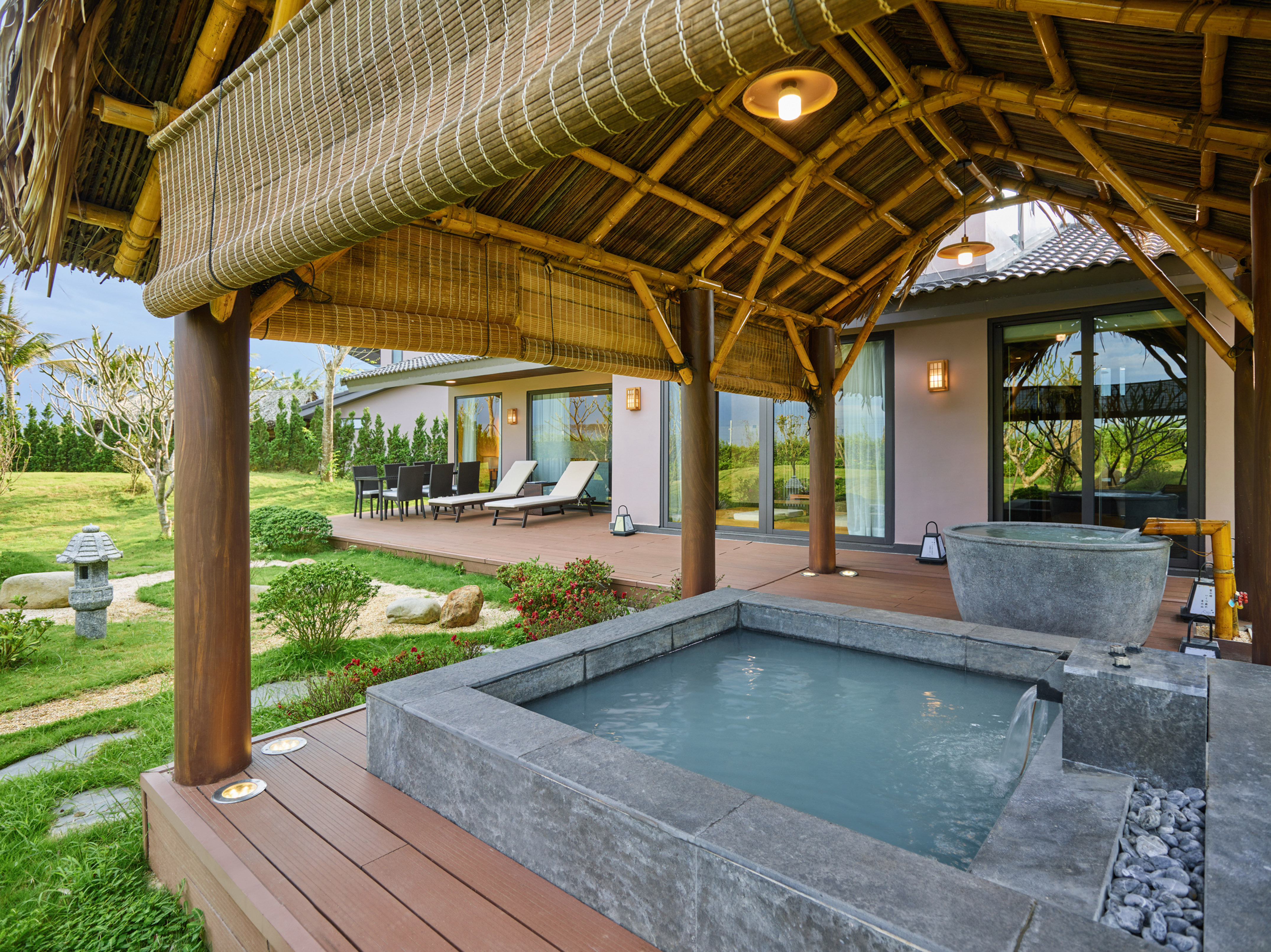 Outdoor spa with stone hot tub, bamboo roof, wooden deck, and green garden.