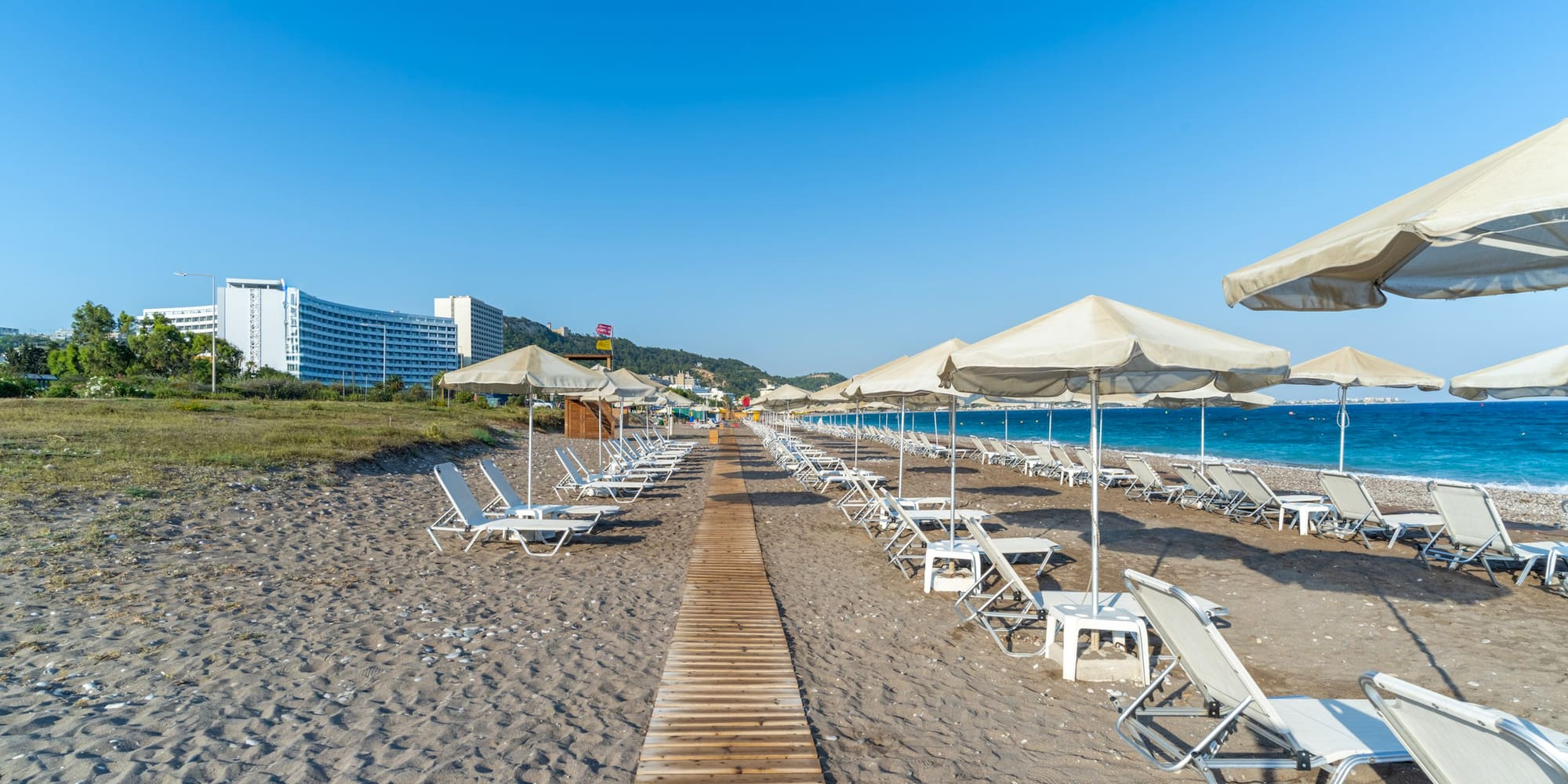 Sandy beach with sun loungers, umbrellas, boardwalk, blue sea, and hotel.