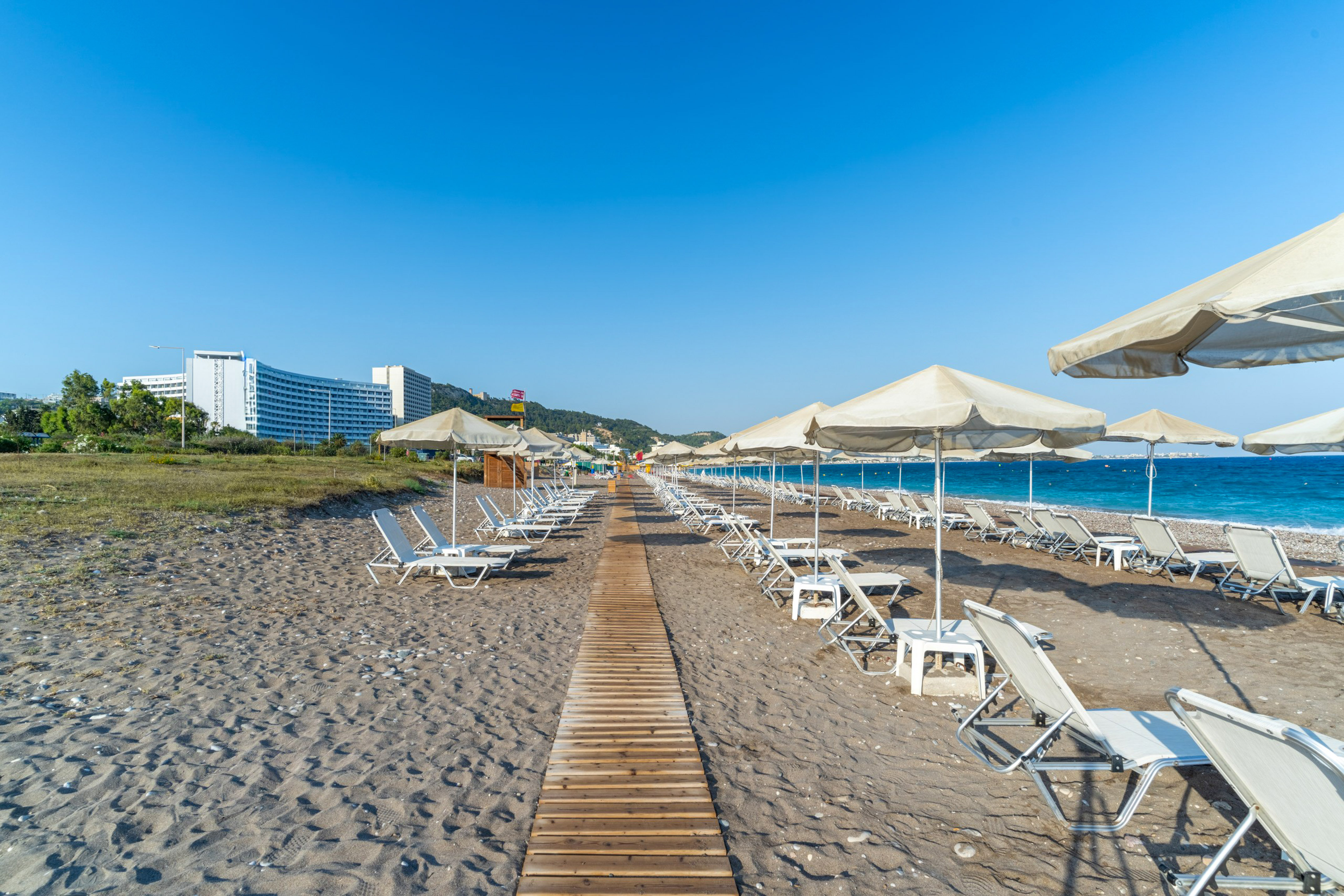 Sandy beach with sun loungers, umbrellas, boardwalk, blue sea, and hotel.