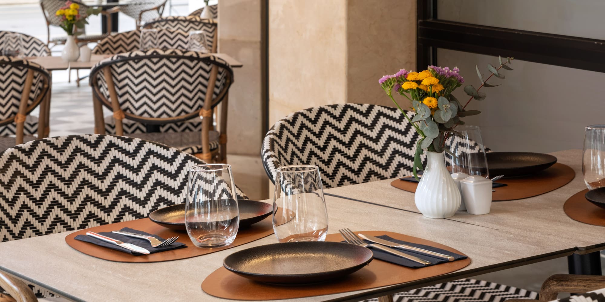 Outdoor restaurant patio with set tables, flowers, and black/white zigzag chairs.