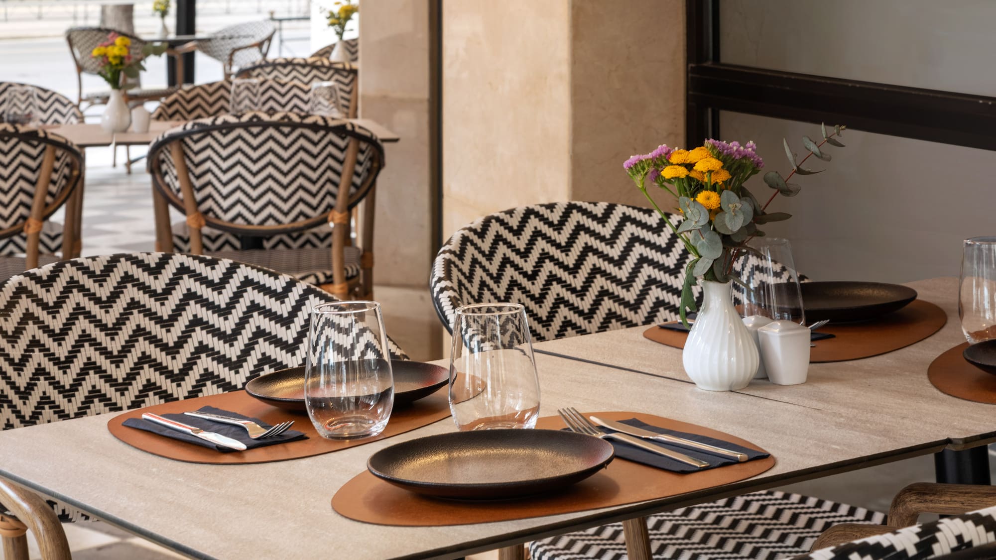 Outdoor restaurant patio with set tables, flowers, and black/white zigzag chairs.