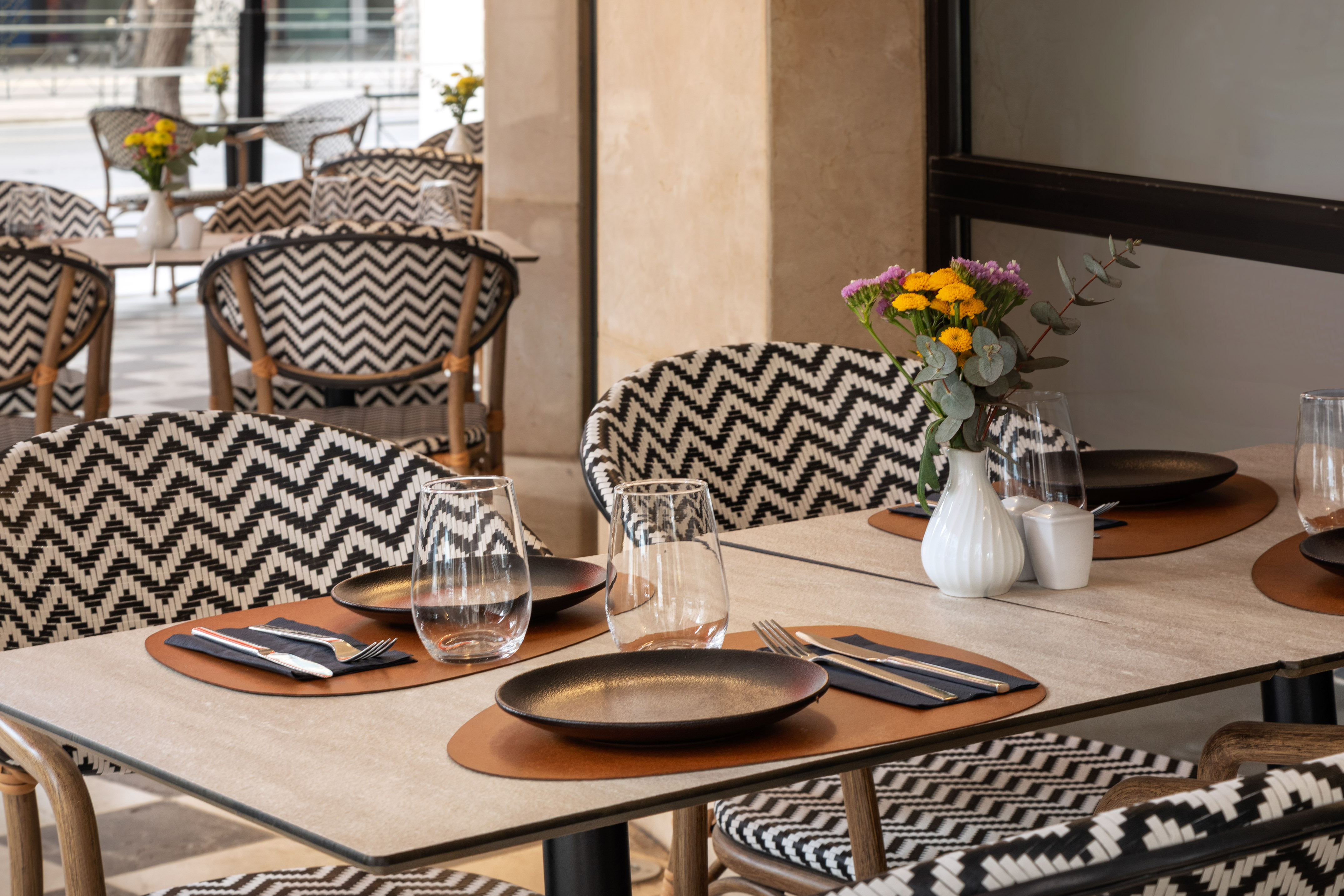 Outdoor restaurant patio with set tables, flowers, and black/white zigzag chairs.
