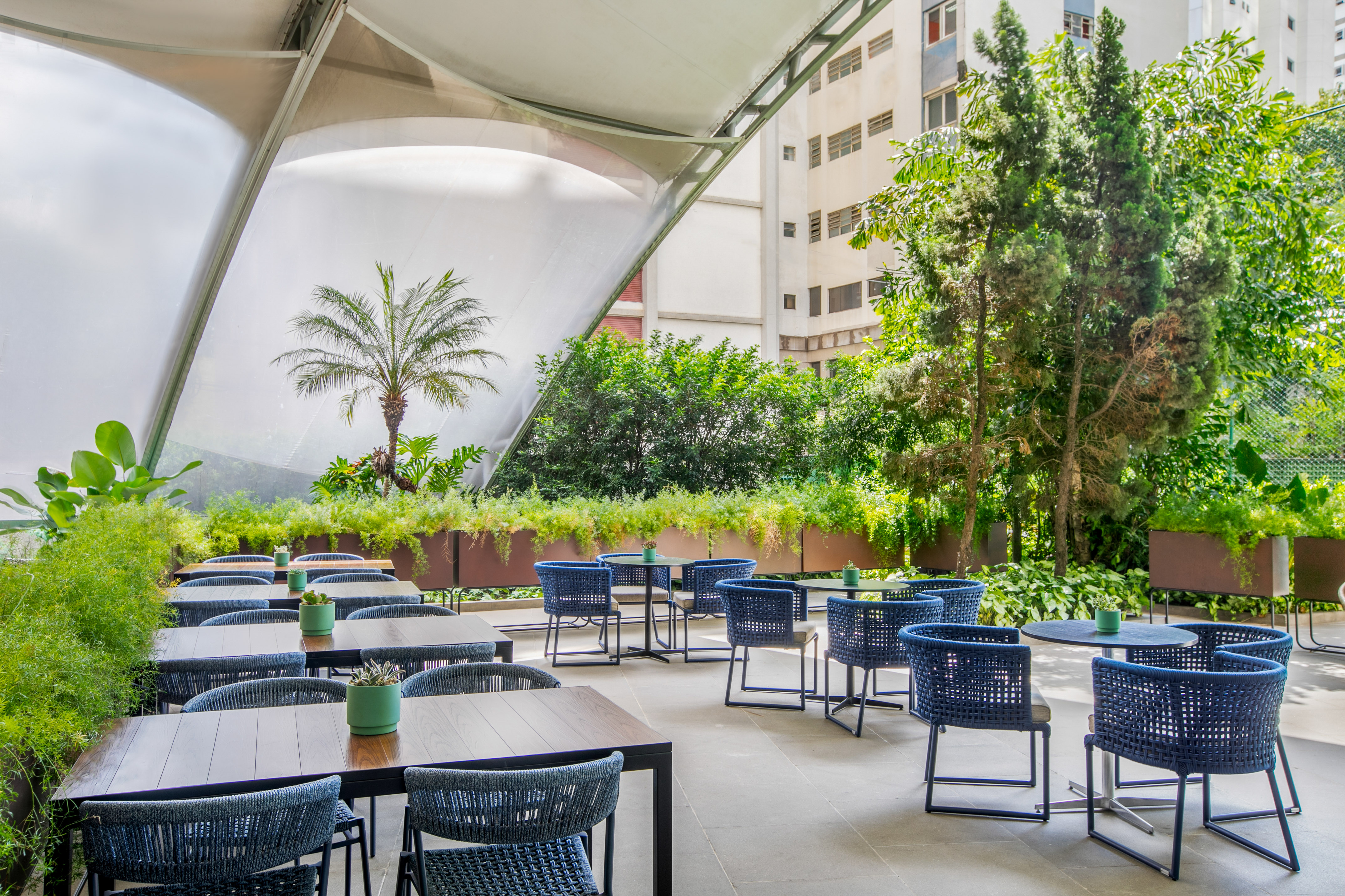 Modern outdoor patio with blue chairs, dark tables, lush plants, and white canopy.
