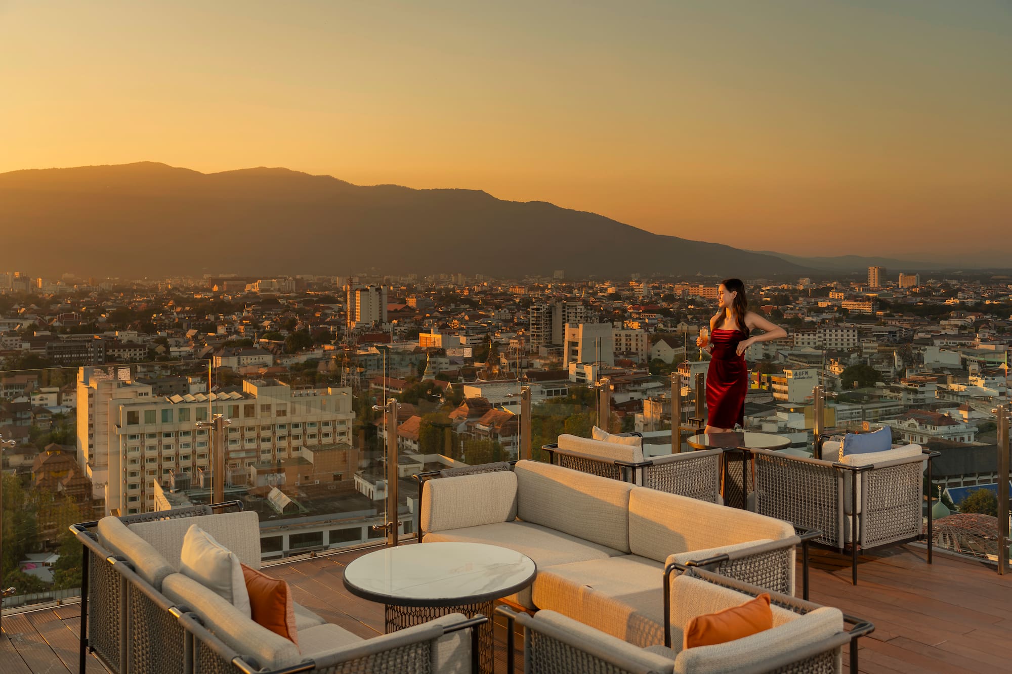 Woman in red dress on rooftop bar, city and mountains at sunset.