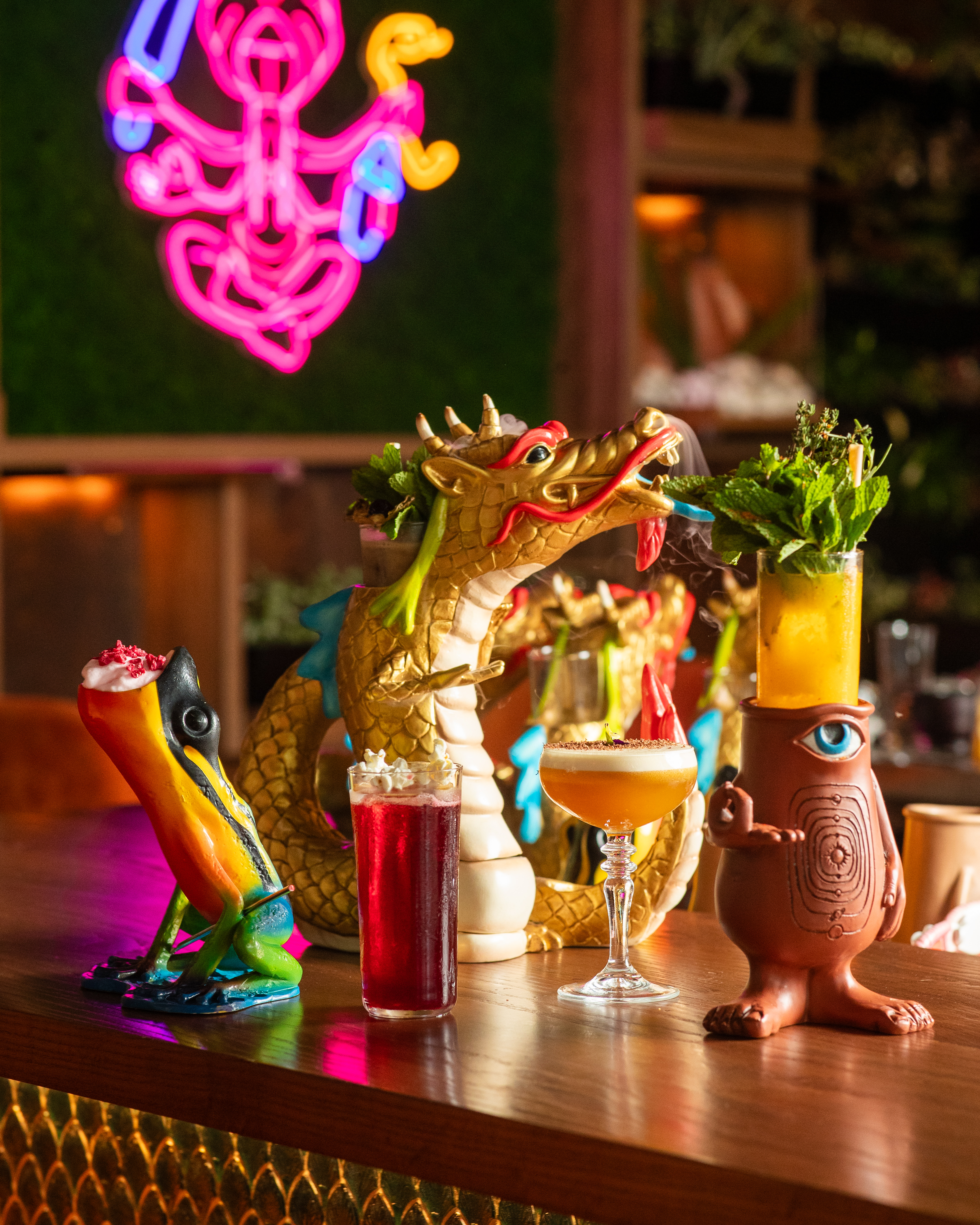 Exotic cocktails in frog, dragon, and eye-character vessels at a vibrant neon bar.