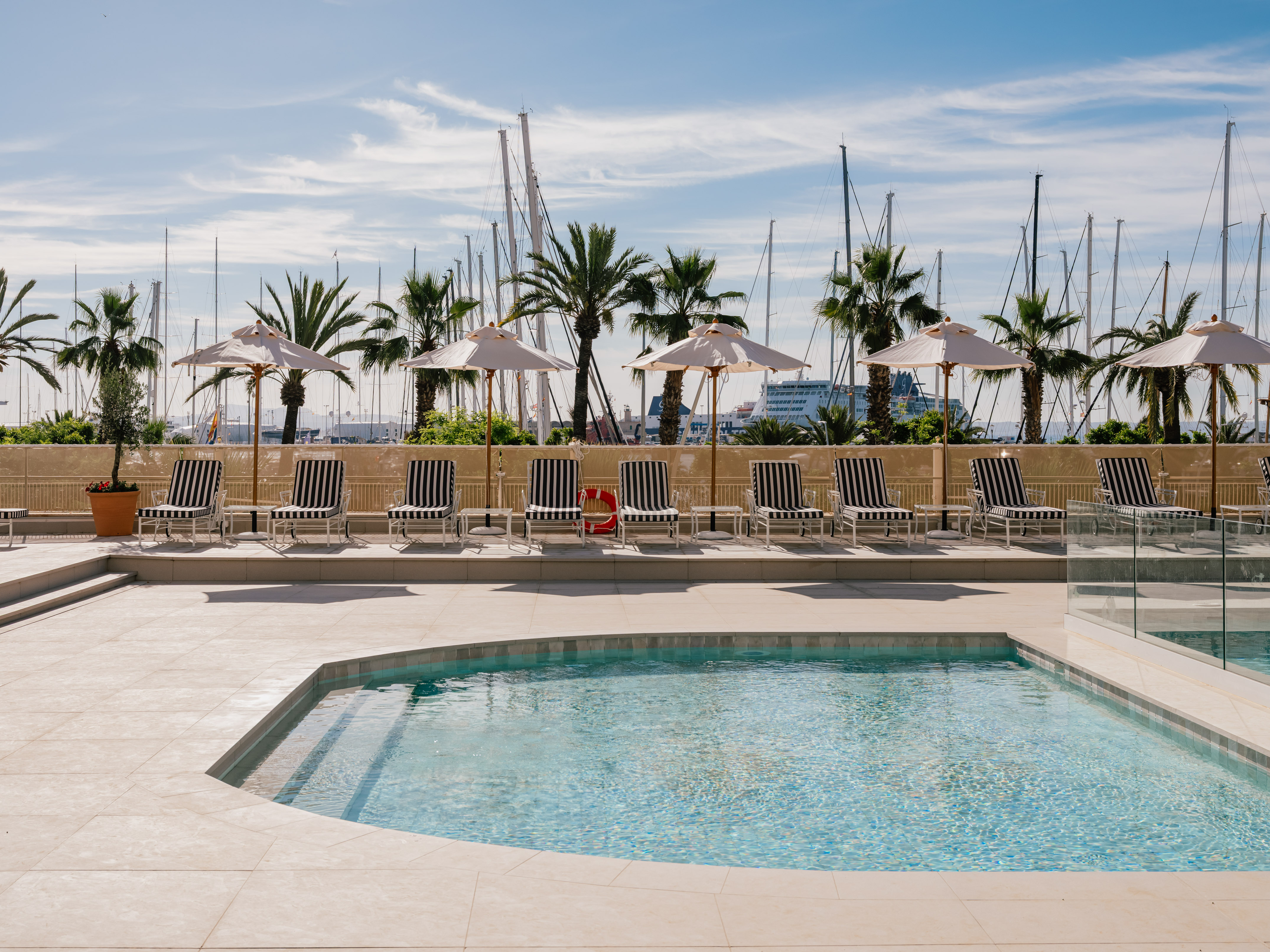 Sunny hotel pool with striped lounge chairs, palm trees, and marina.
