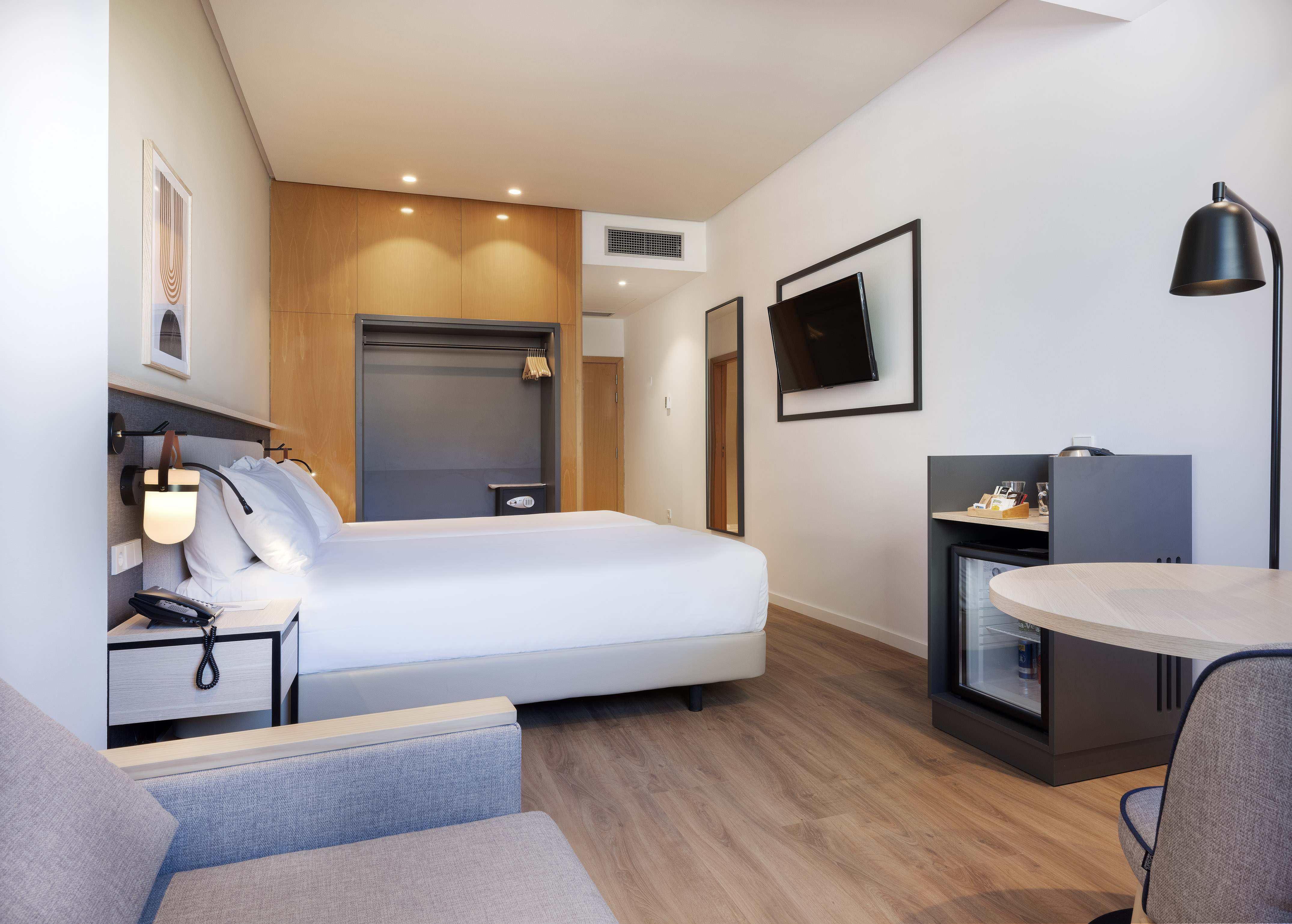 Modern hotel guest room with white bed, wall-mounted TV, work desk, and minibar.