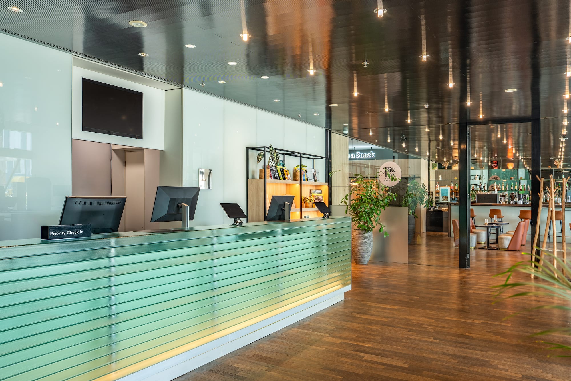 Modern hotel lobby with green reception desk, check-in monitors, and bar.
