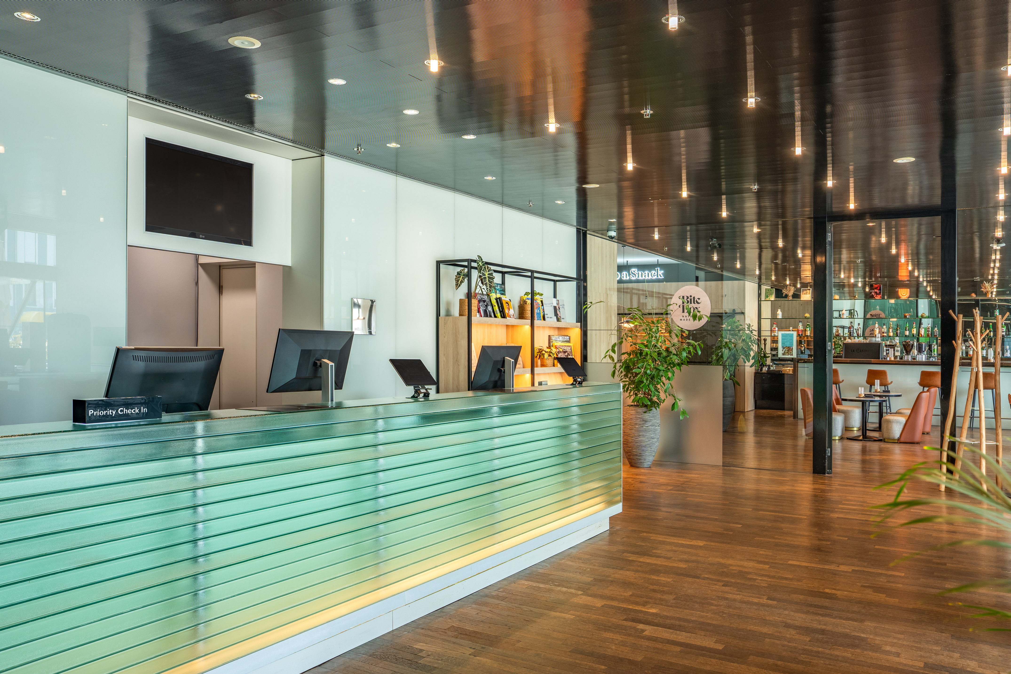 Modern hotel lobby with green reception desk, check-in monitors, and bar.
