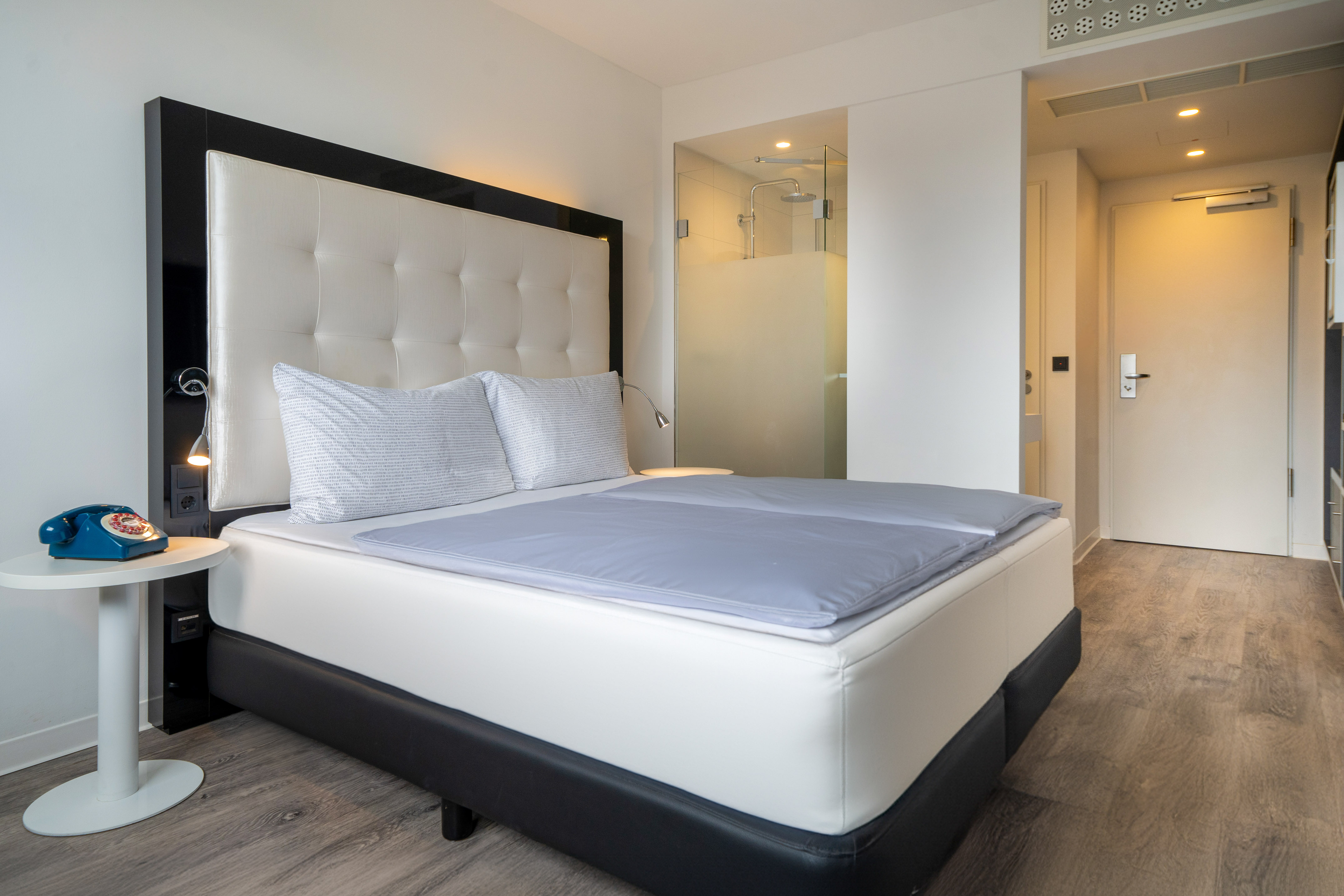 Modern hotel room with large bed, blue rotary phone, and glass shower.