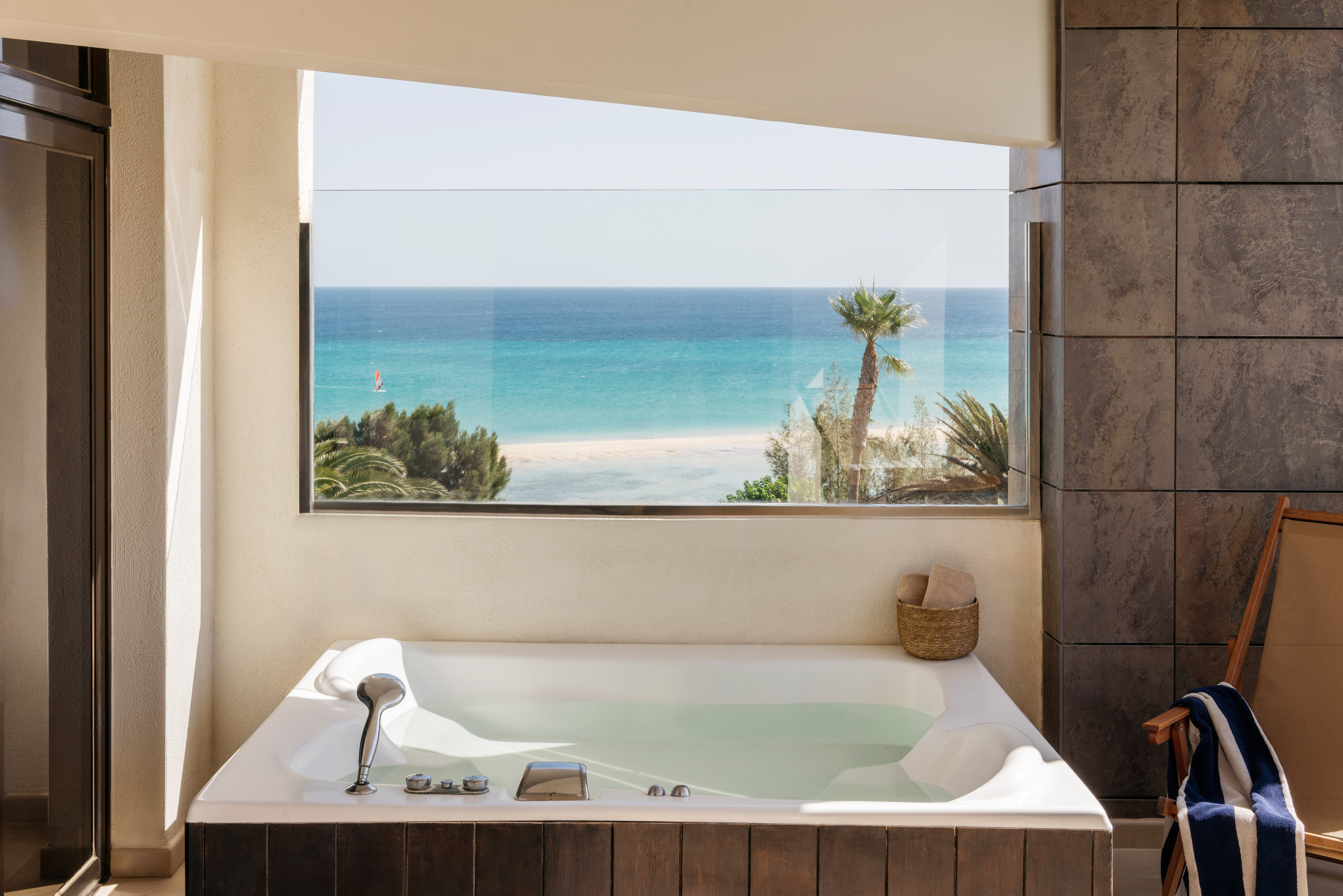 White jacuzzi with ocean view, tropical beach, palm trees, and windsurfer.