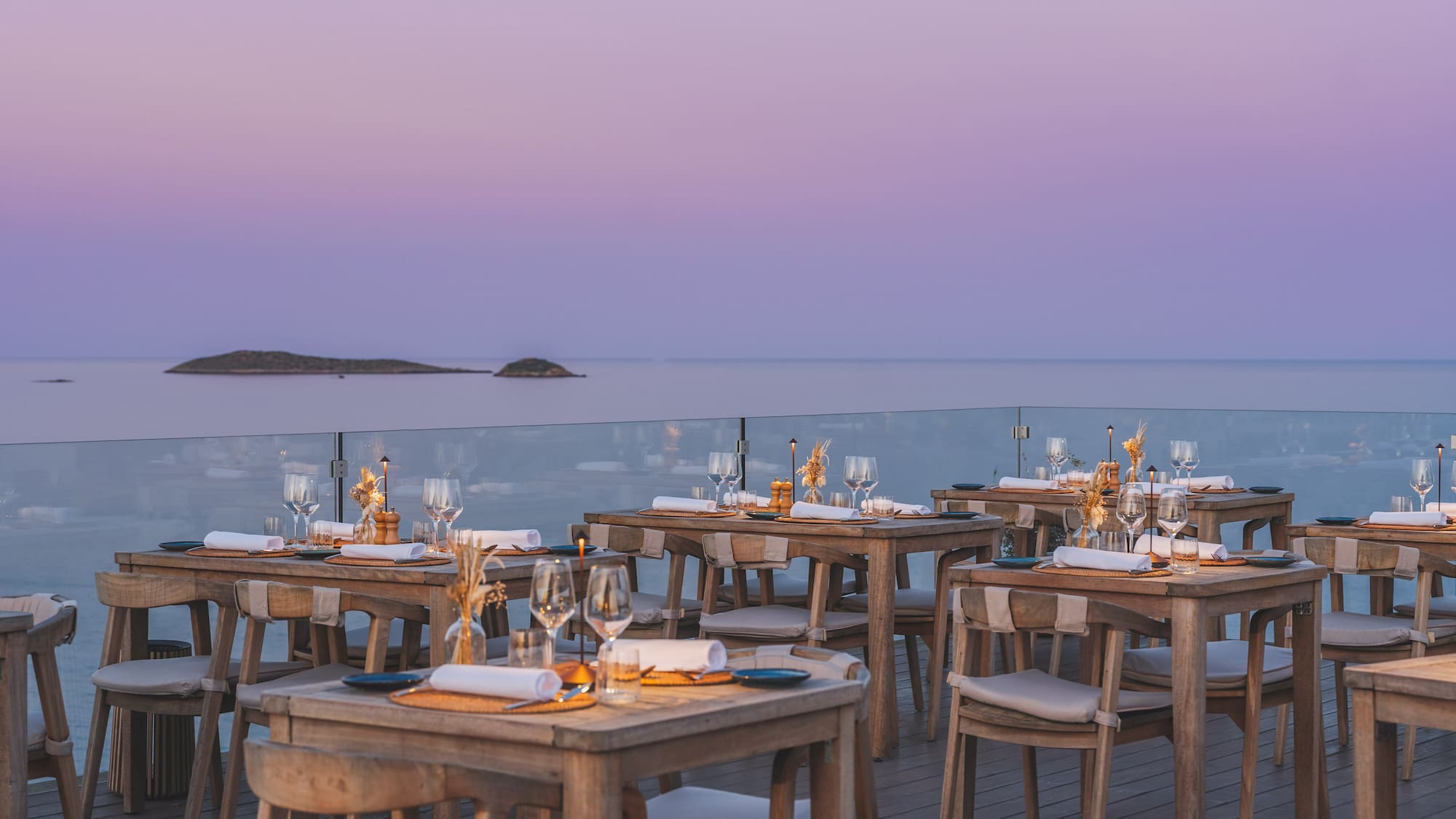 Outdoor dining terrace with wooden tables overlooking the ocean at sunset.