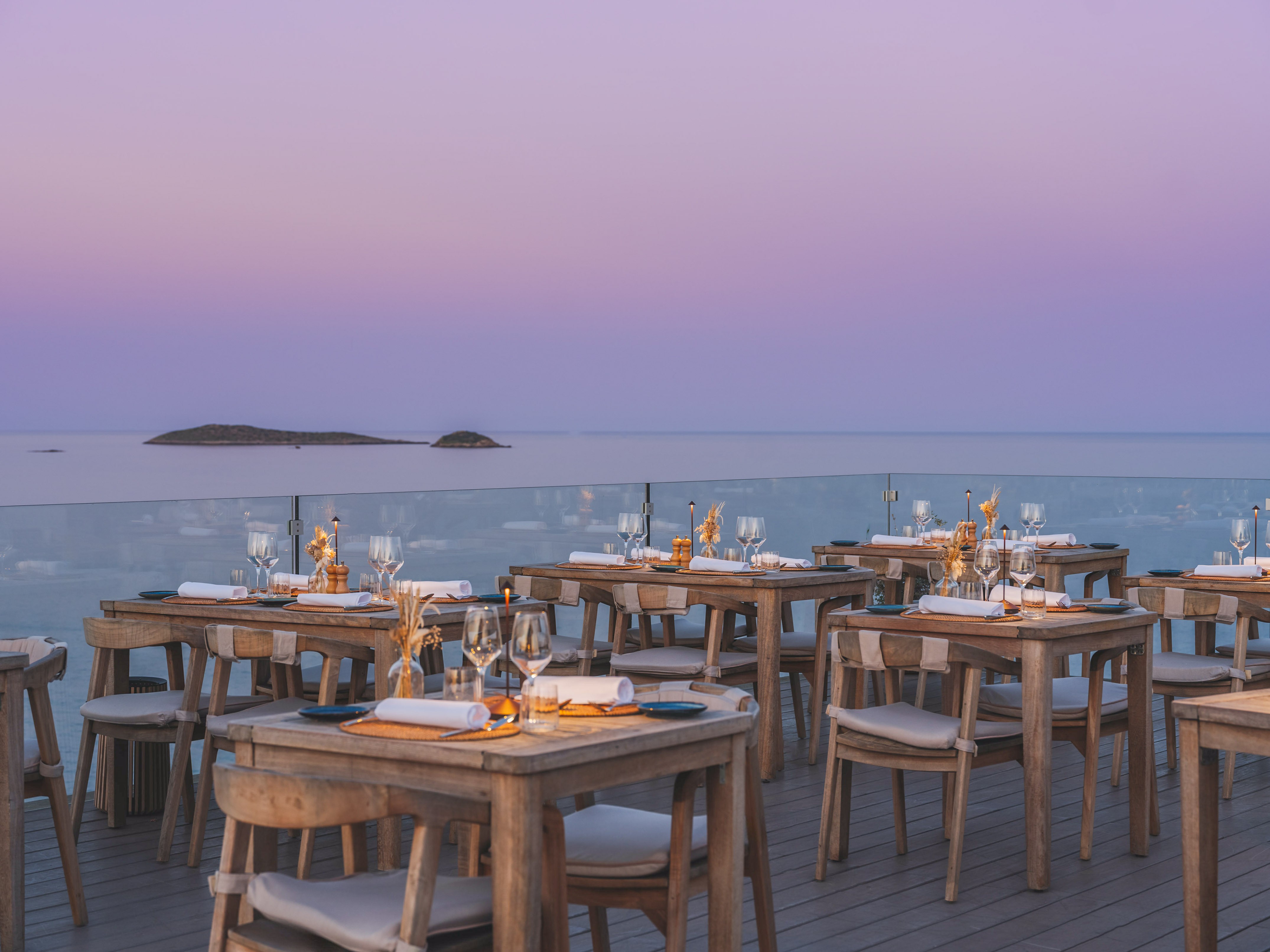 Outdoor dining terrace with wooden tables overlooking the ocean at sunset.