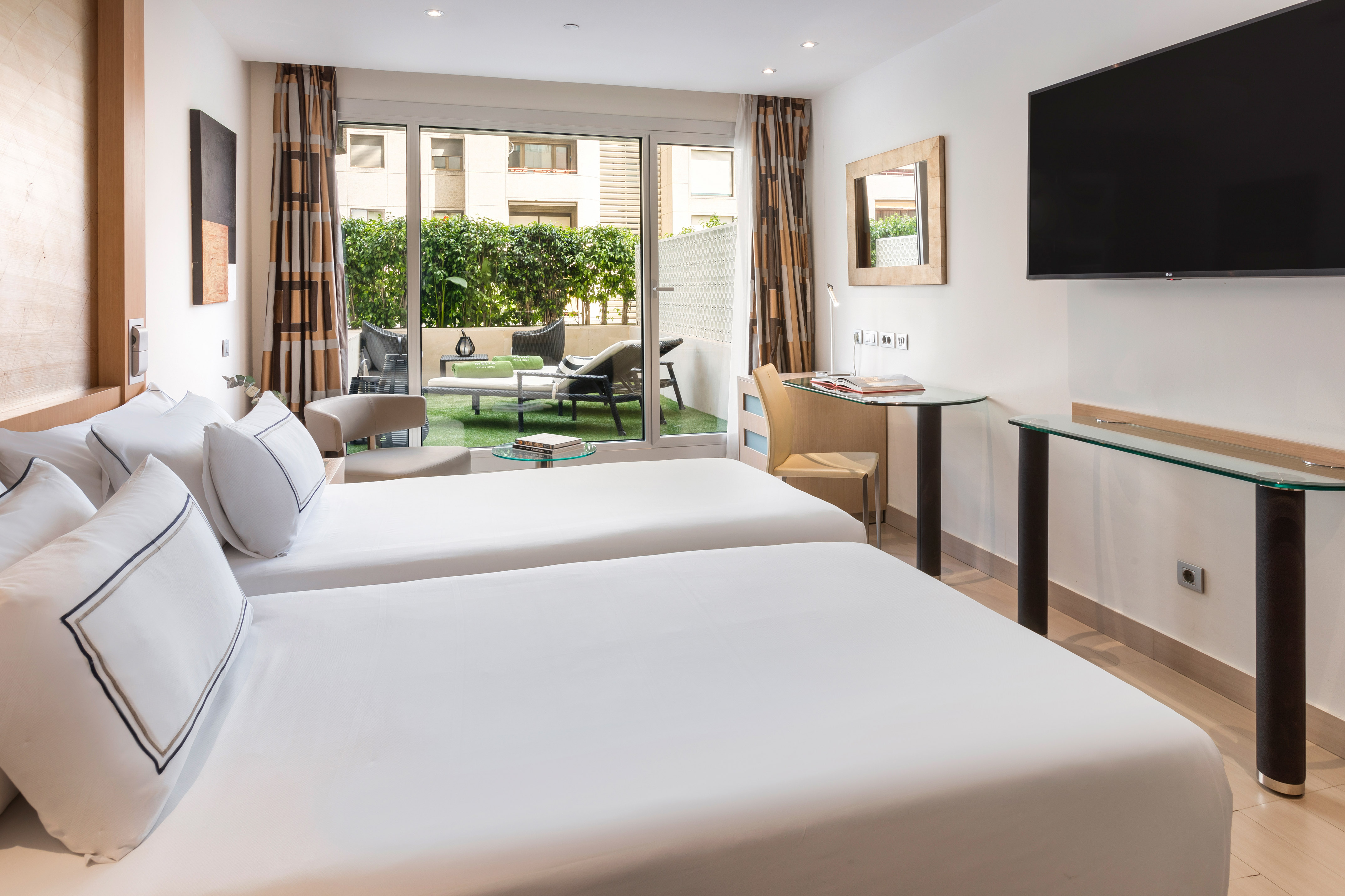 Bright hotel room with twin beds and private terrace with lounge chairs.