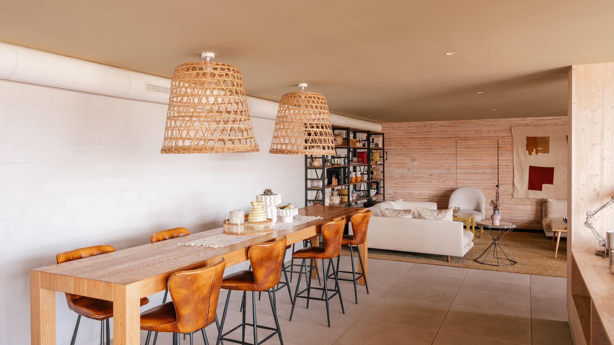 Modern open-plan dining and living room with wooden table, leather stools, rattan lights.