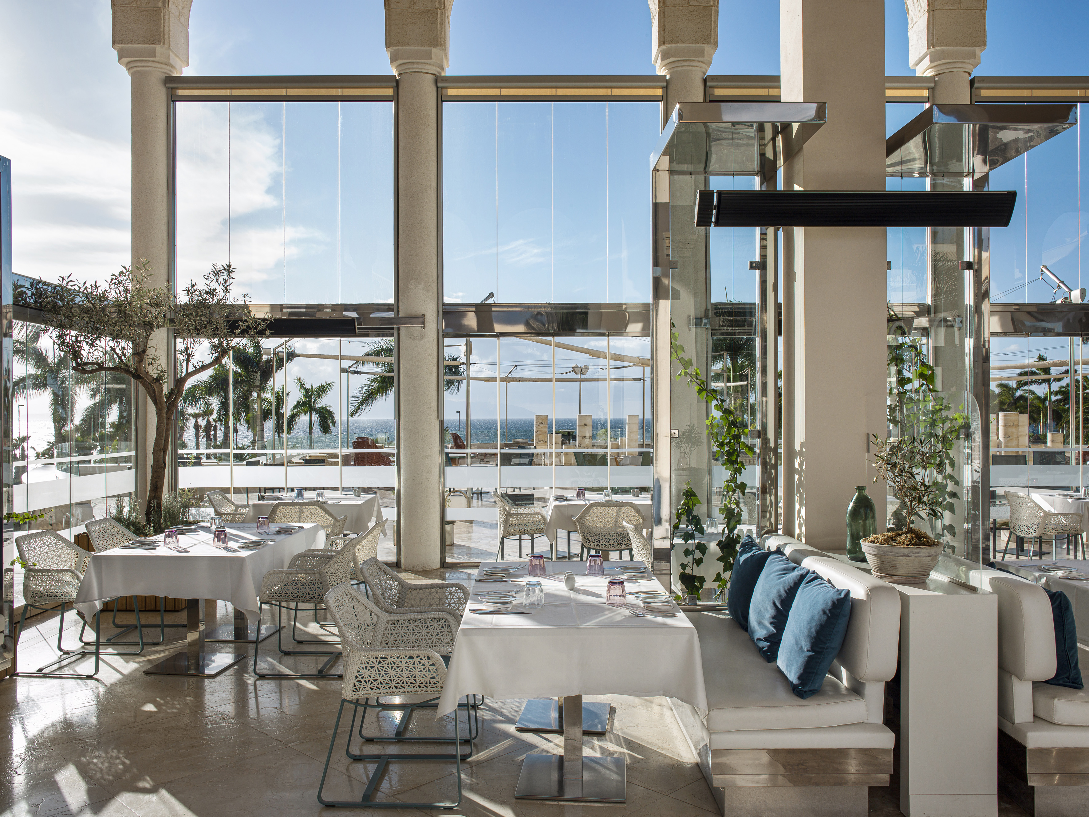 Modern restaurant with ocean view, white dining tables, and tropical plants.
