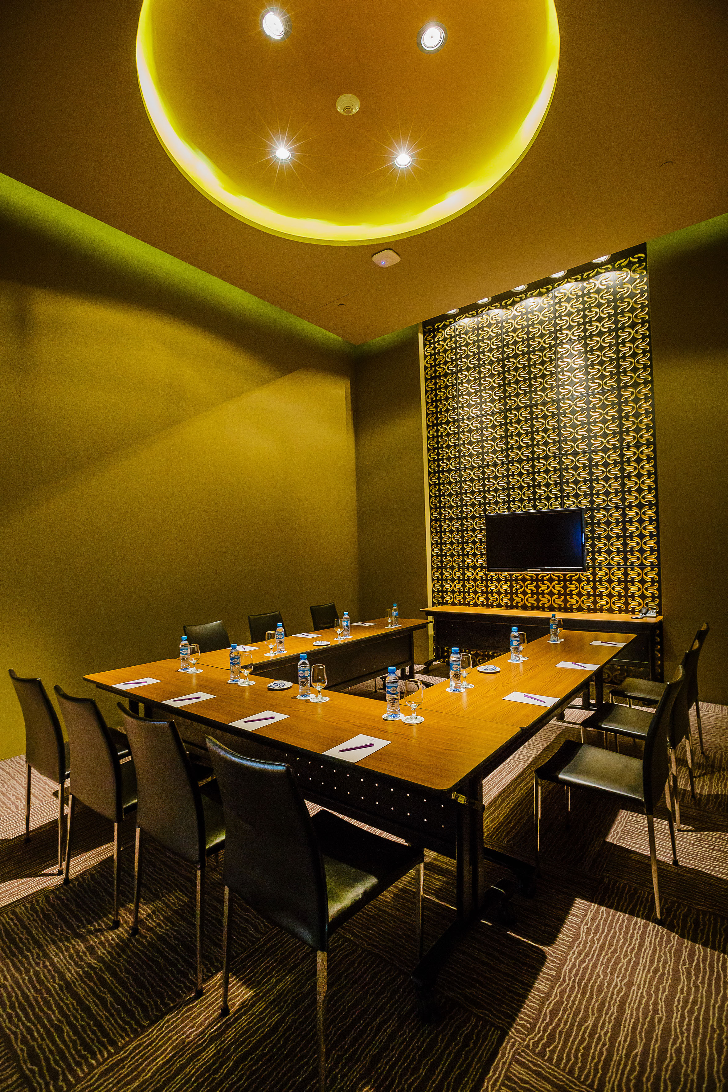Modern conference room with U-shaped table, olive walls, TV, and warm lighting.