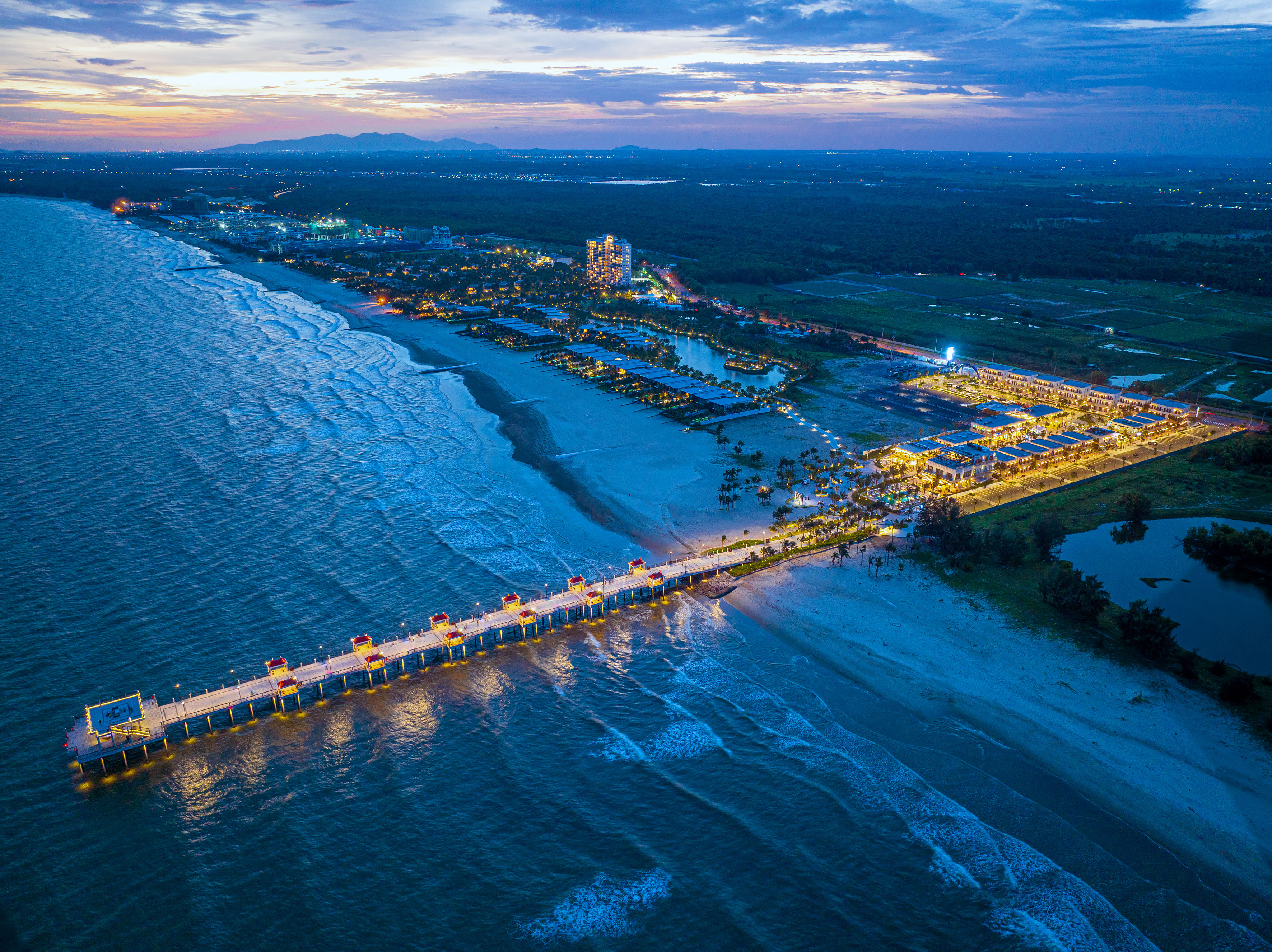 Aerial view: illuminated coastal resort, pier, and beach at dusk.