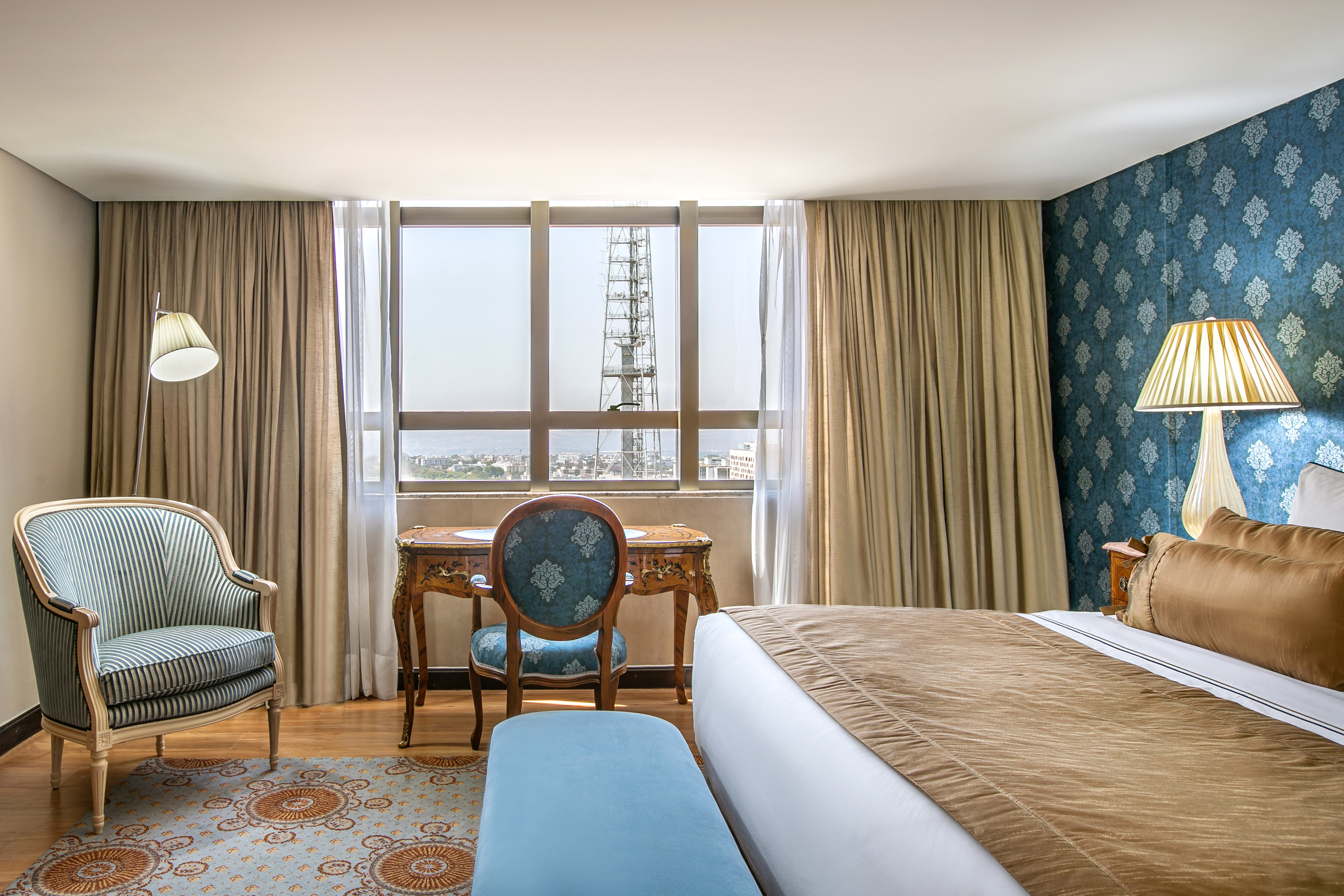 Elegant hotel room: king bed, ornate desk, armchair, city window view.
