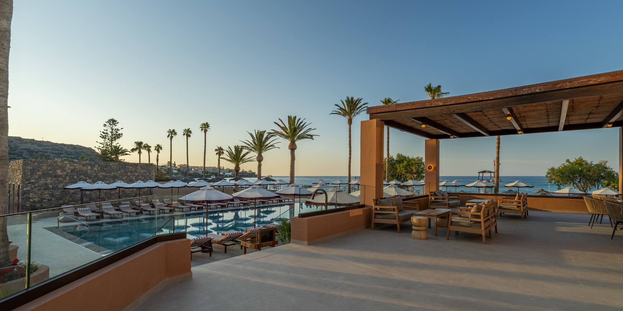 Luxury resort pool, outdoor lounge, ocean view, and palm trees at sunset.