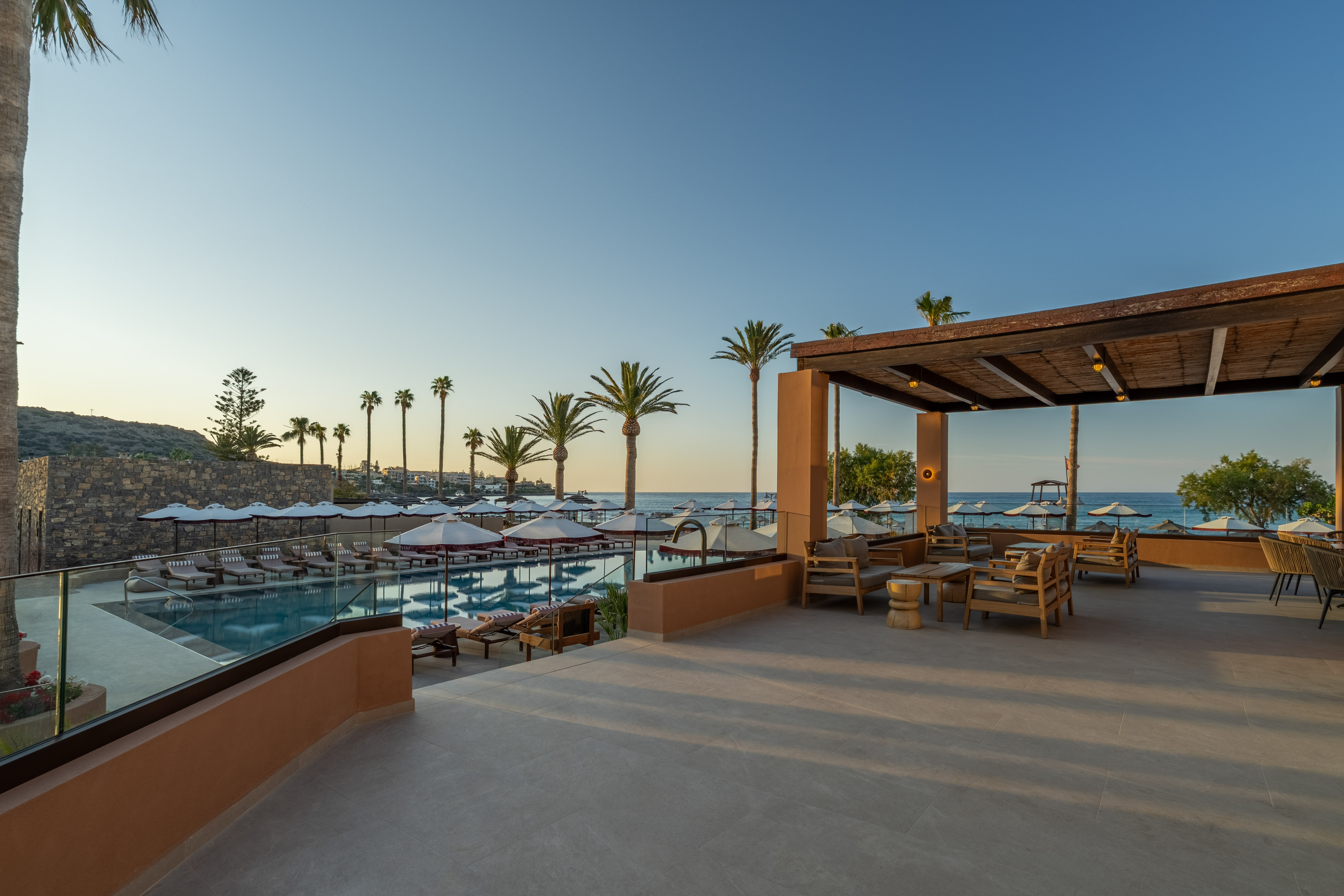 Luxury resort pool, outdoor lounge, ocean view, and palm trees at sunset.
