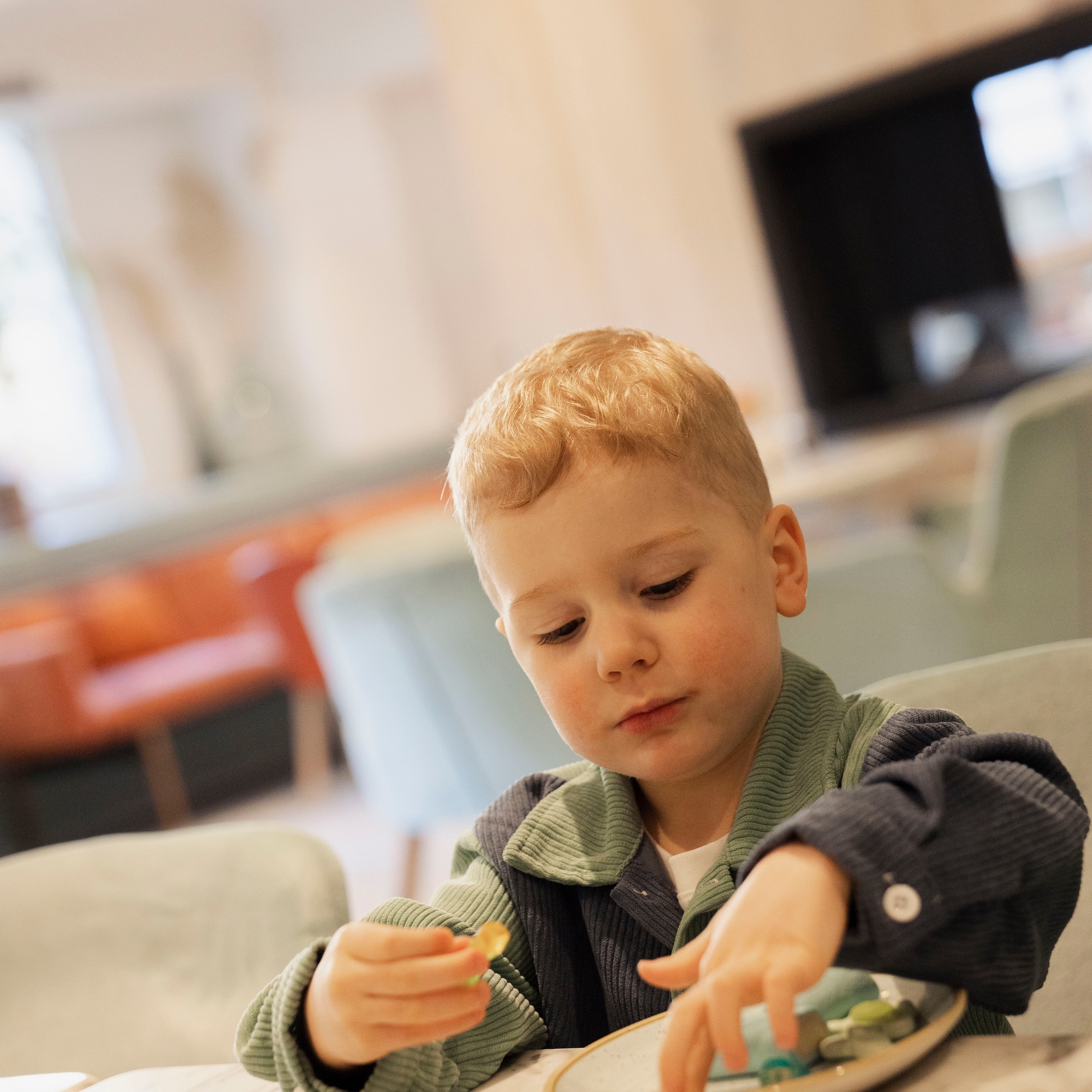 Young blonde boy playing with candies on a plate at a cafe table.